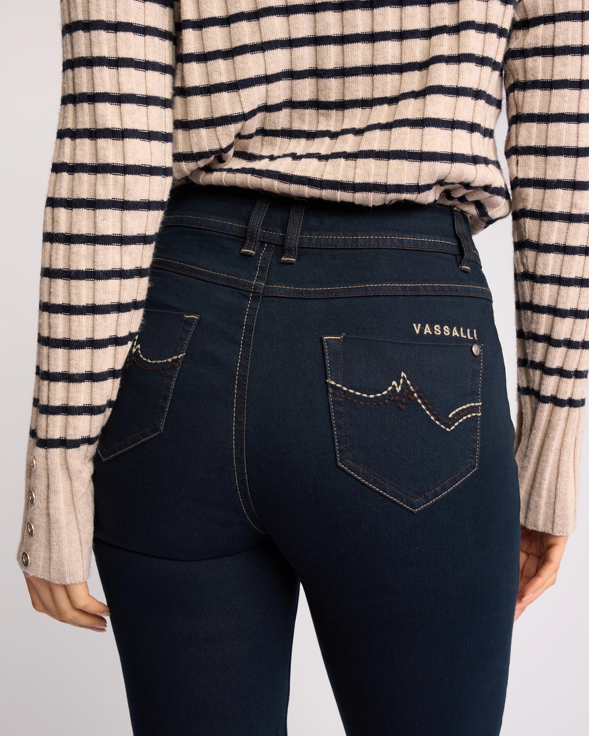 Wearing Vassalli’s Pace Jean in Indigo Denim, featuring stretch fabric and back pocket embroidery, paired with a beige striped long-sleeve sweater. The image highlights the lower back and waist.