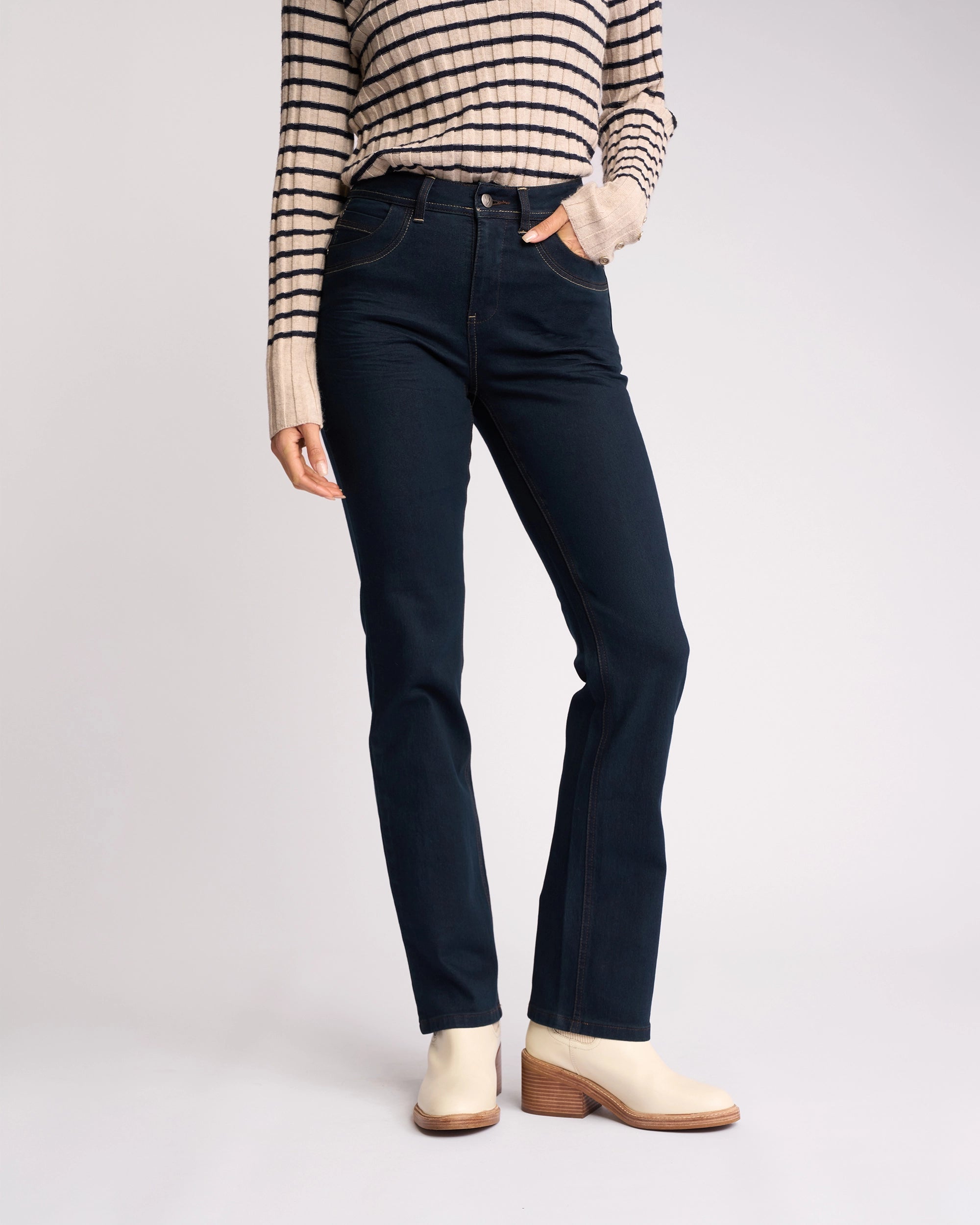 A person wears the Vassalli Pace Jean in Indigo Denim, a beige and black striped long-sleeve top, and off-white chunky-heeled ankle boots, standing with one hand in their pocket against a plain background.