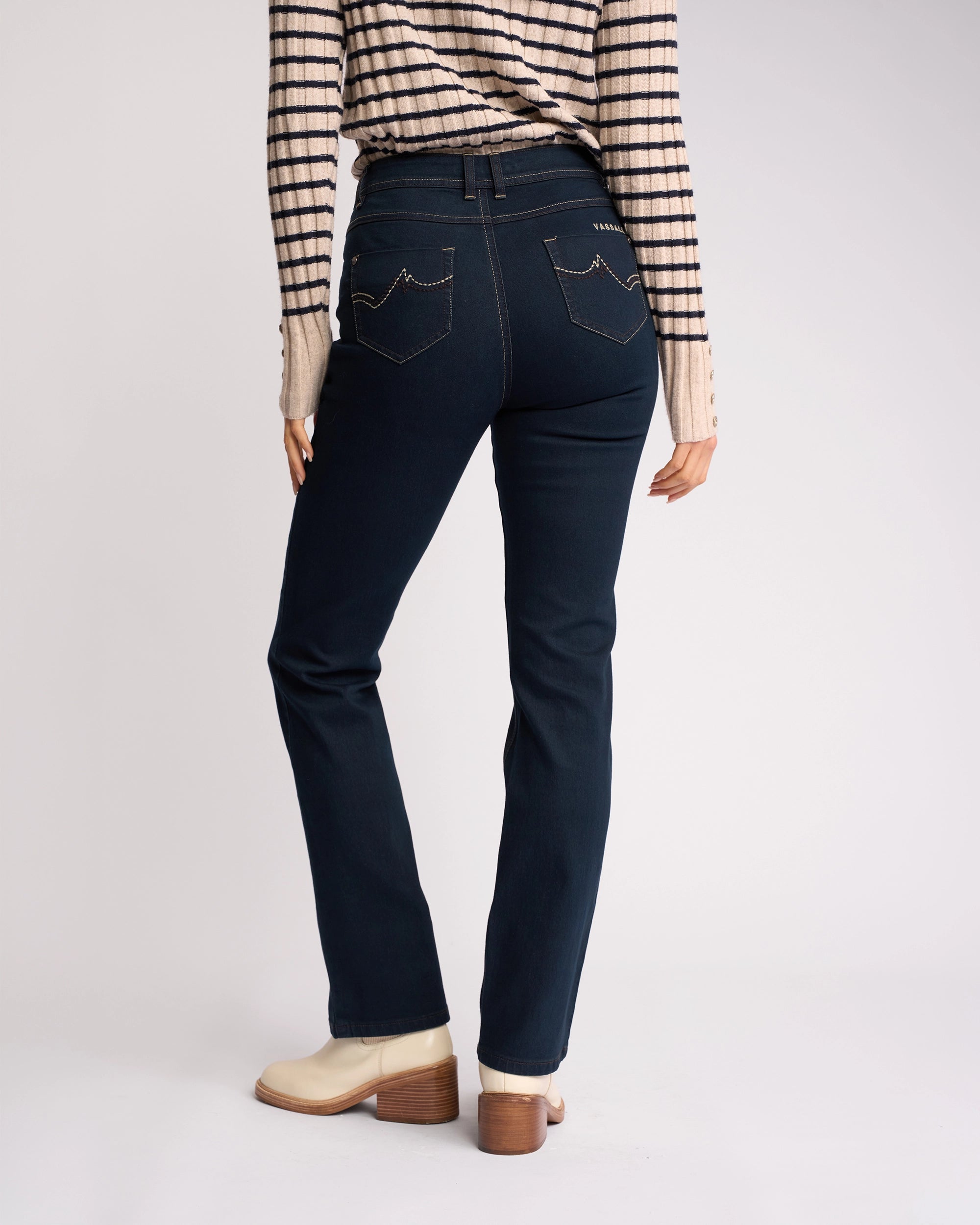 A person wearing Vassalli Pace Jean in indigo denim, a striped long-sleeve shirt, and light ankle boots stands with their back to the camera against a plain background.