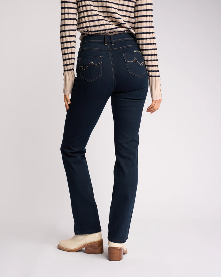 A person wearing Vassalli Pace Jean in indigo denim, a striped long-sleeve shirt, and light ankle boots stands with their back to the camera against a plain background.