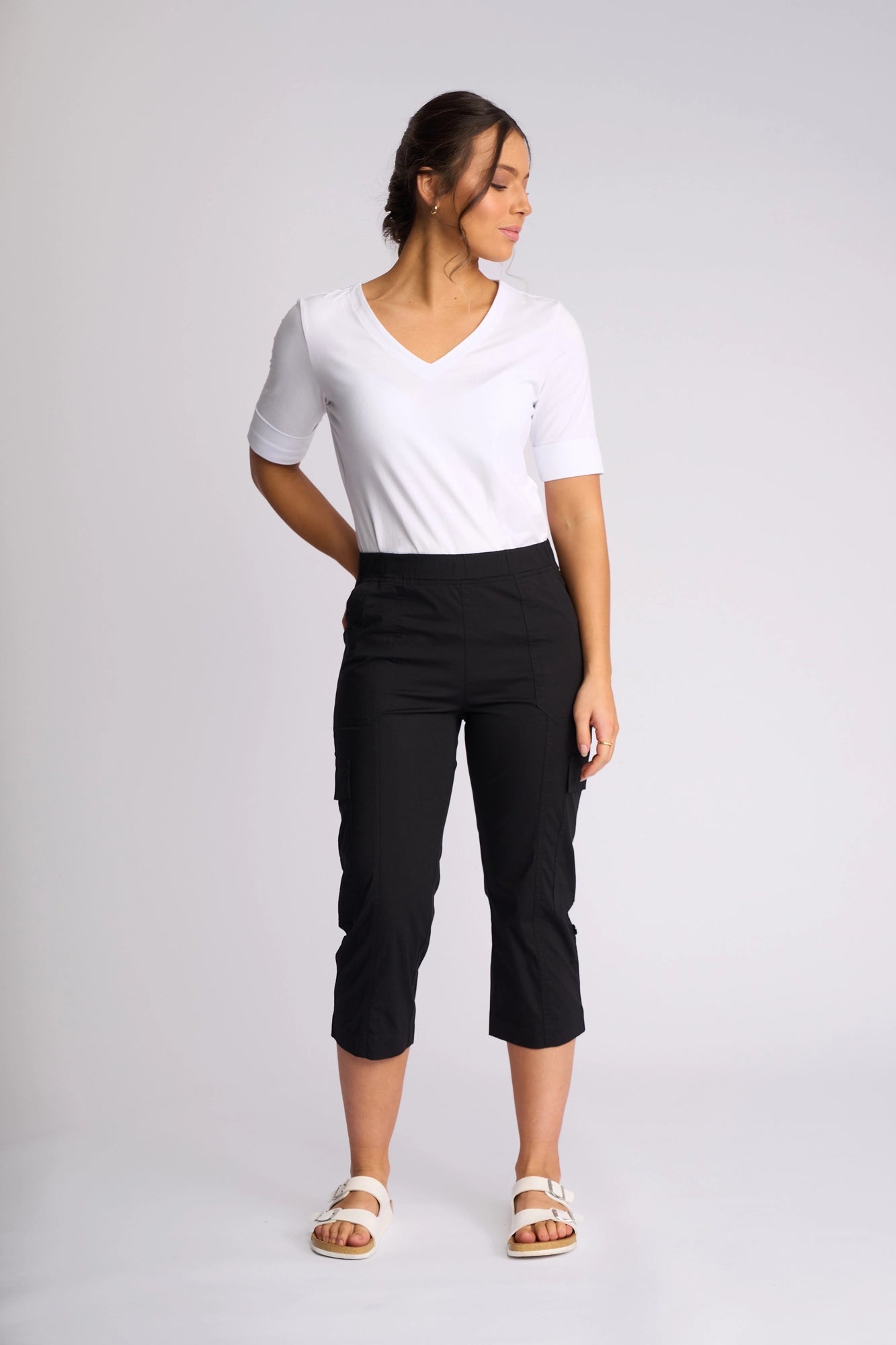 A woman stands against a plain backdrop, wearing a white V-neck top, the Vassalli Wave Pant in black with adjustable hems, and white sandals. She faces slightly to the side with her hands behind her back.