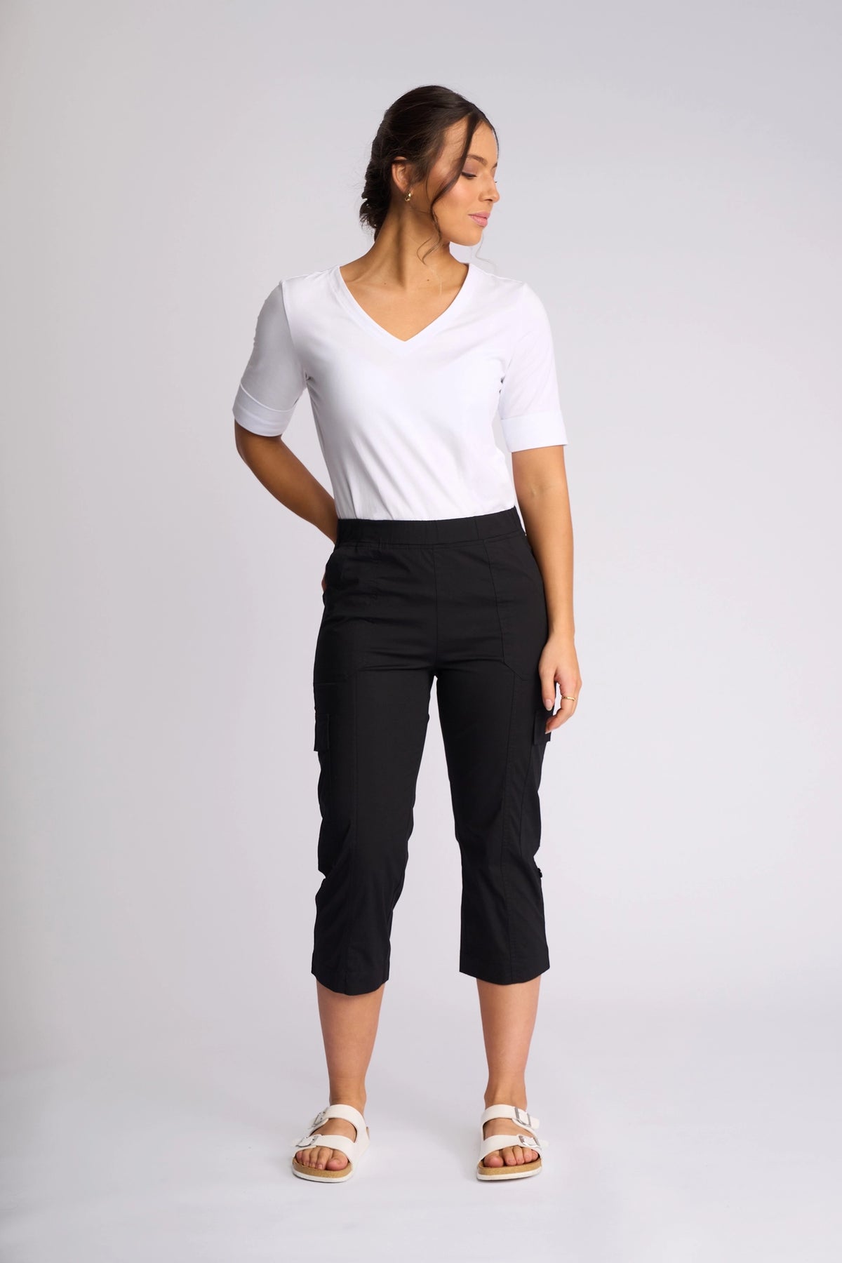 A woman stands against a plain backdrop, wearing a white V-neck top, the Vassalli Wave Pant in black with adjustable hems, and white sandals. She faces slightly to the side with her hands behind her back.