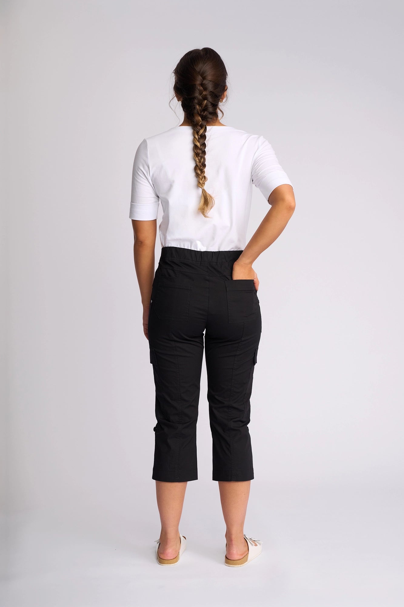 A woman with a long braid, dressed in a white shirt, Vassalli’s Wave Pant in Black featuring adjustable hems, and sandals, stands facing away from the camera against a plain light background.