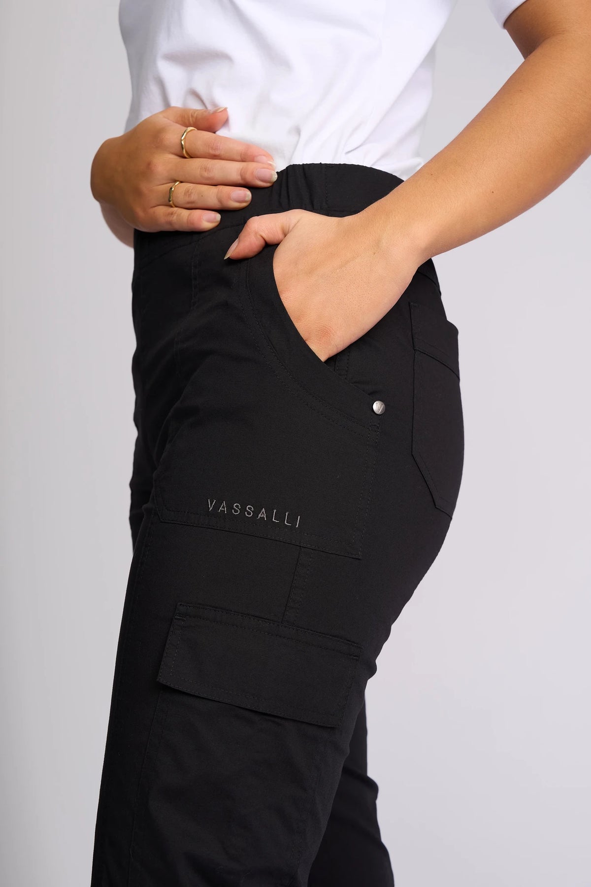 A person wearing Vassalli’s Wave Pant in Black, featuring adjustable hems, paired with a white shirt; one hand is in their pocket while the other rests on their waist, displaying gold rings on their fingers.