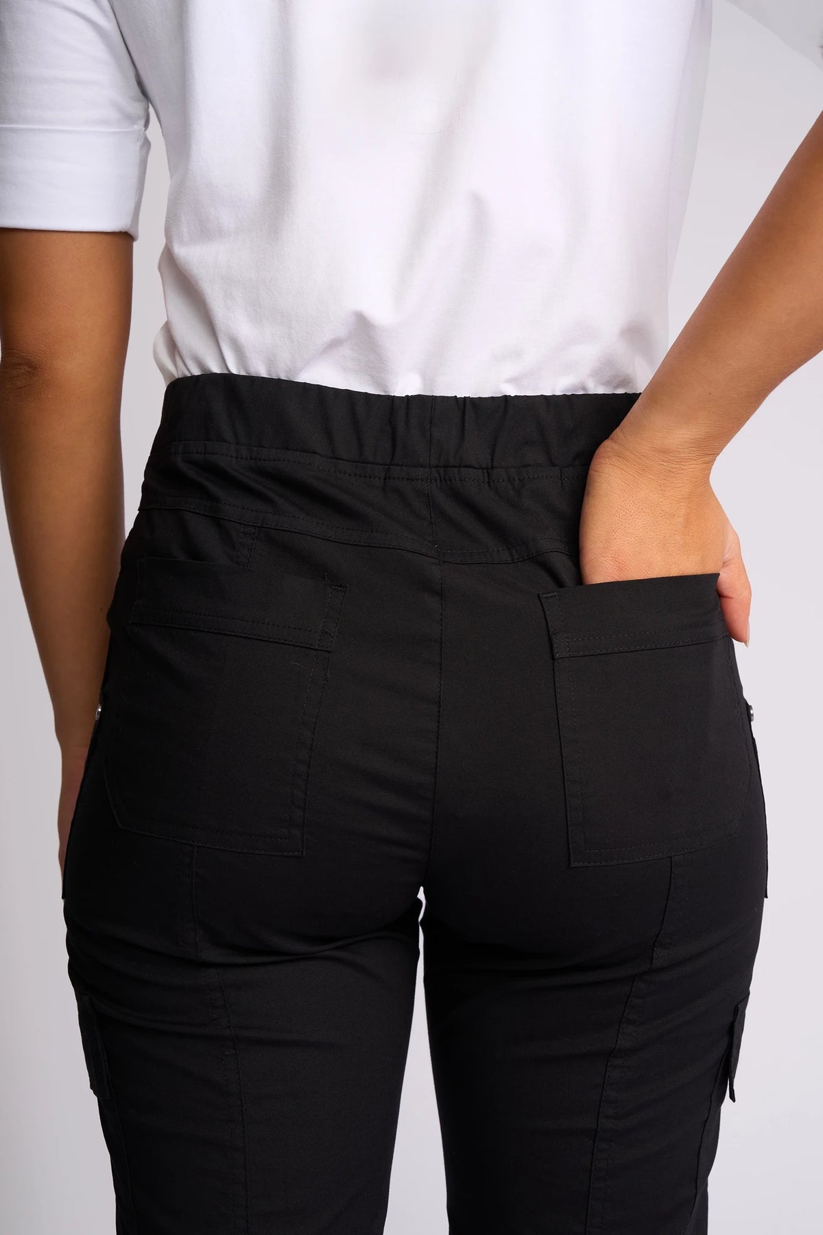 A person is seen from behind wearing the Vassalli Wave Pant in black, paired with a white shirt. Their right hand is slipped into the back pocket of the pants, drawing attention to the waist and upper legs and highlighting the stretch cotton fabric and adjustable hems for a comfortable, tailored fit.