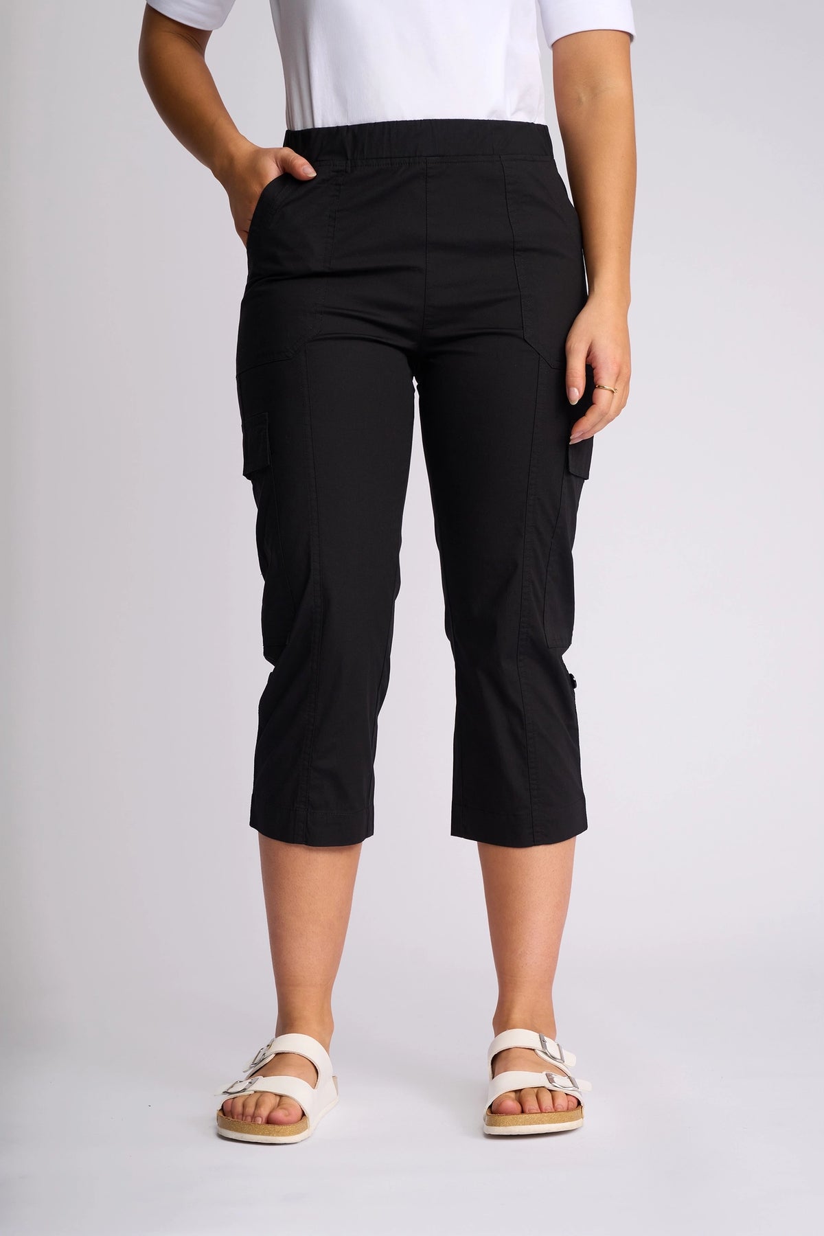 A person wearing the Vassalli Wave Pant in black—cropped poplin cargo pants with adjustable hems—paired with a white short-sleeve top and white sandals, standing against a plain background.