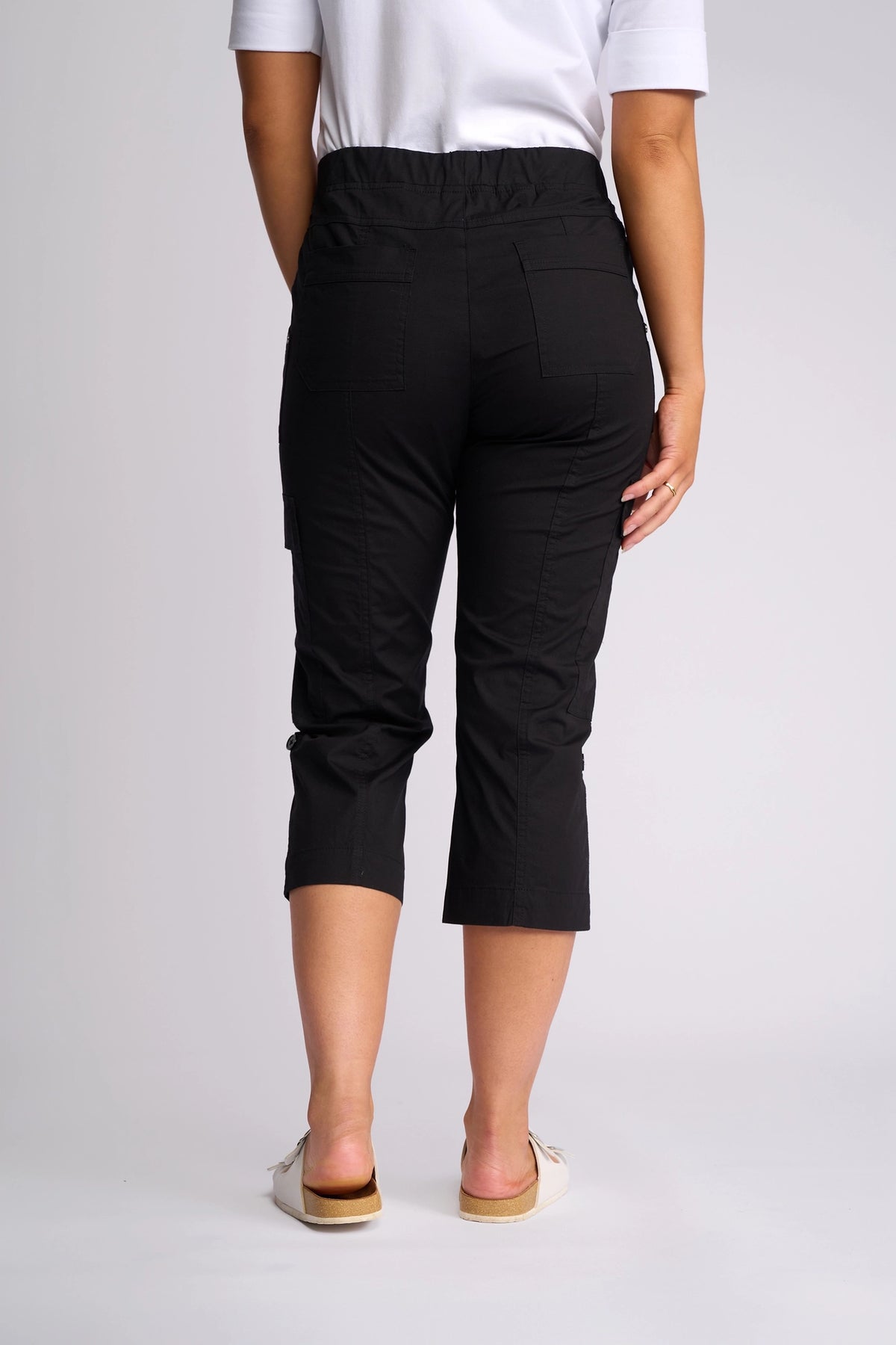 A person wearing a white shirt, the Vassalli Wave Pant in black with adjustable hems, and white slip-on shoes is standing with their back to the camera against a plain gray background.