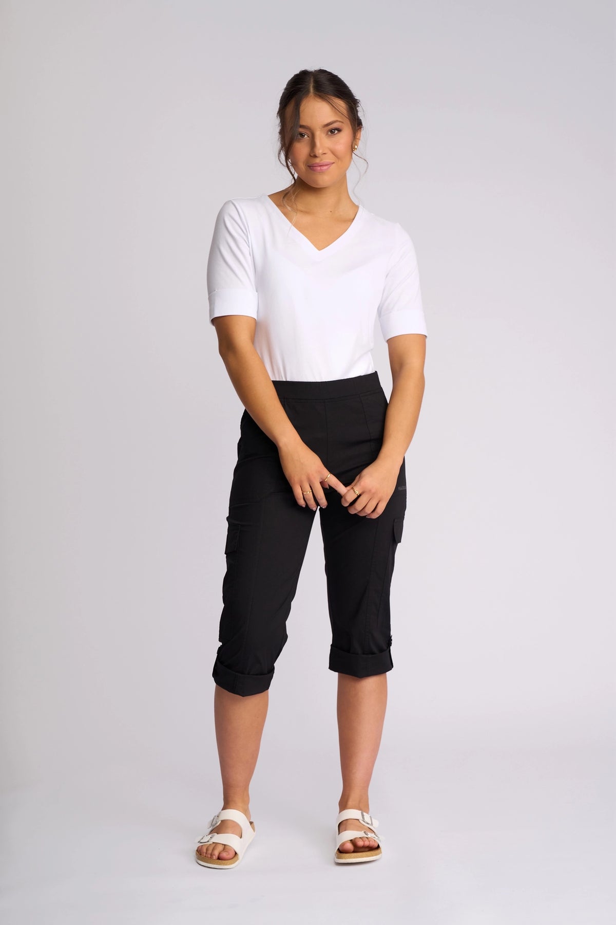 A woman stands against a plain background, wearing a white short-sleeve top and the Vassalli Wave Pant in black, which features adjustable hems. She completes her outfit with white sandals, has her hands clasped, and smiles softly at the camera.