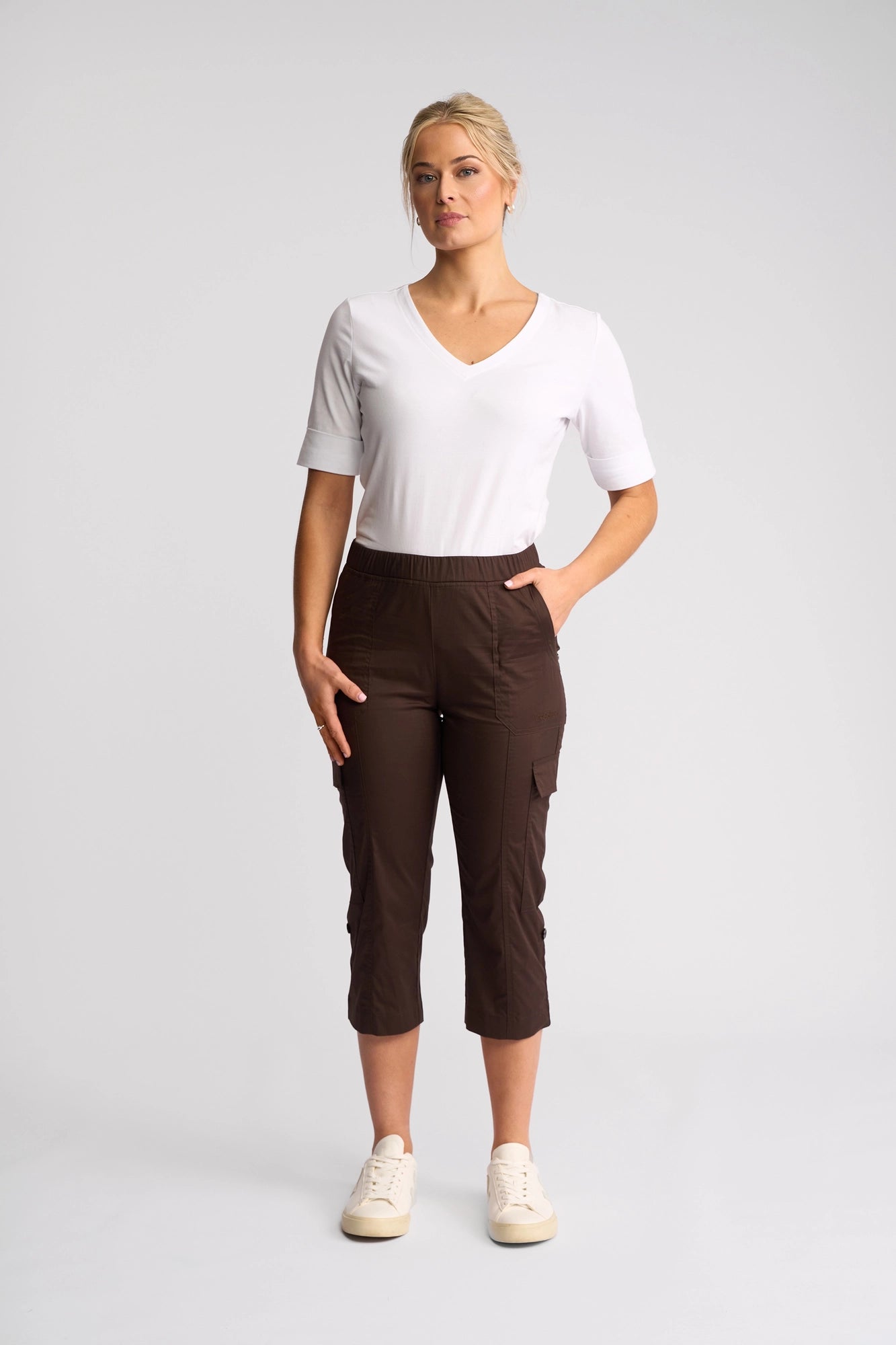 A woman stands facing forward against a plain light background, wearing the Vassalli Wave Pant in Chocolate with an adjustable hem, paired with a white short-sleeve V-neck shirt and cream sneakers, one hand in her pocket.