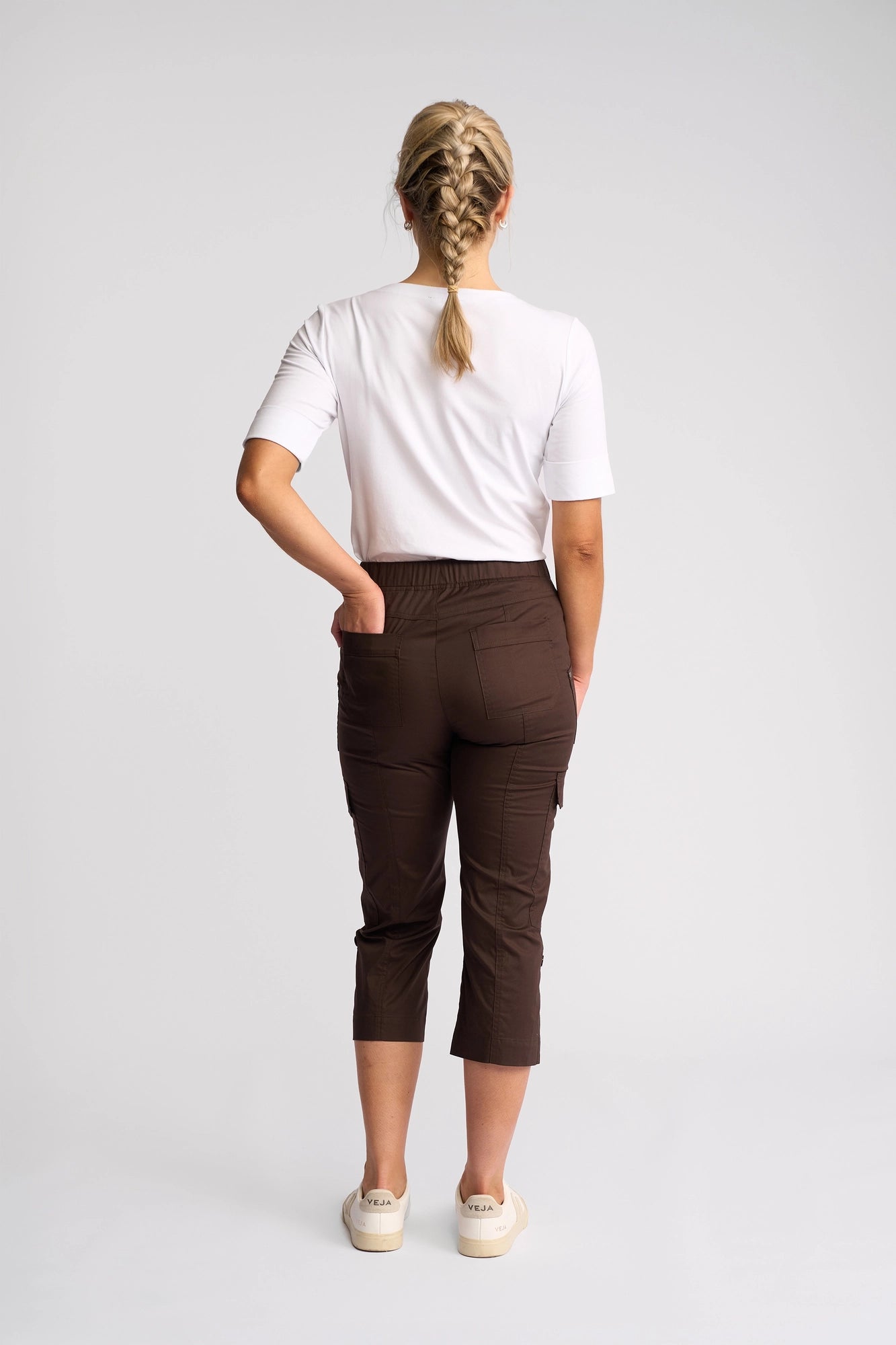 A person with blonde hair in a braid wears a white shirt, the Vassalli Wave Pant in Chocolate, and white shoes, standing facing away with one hand on their hip against a plain light background.