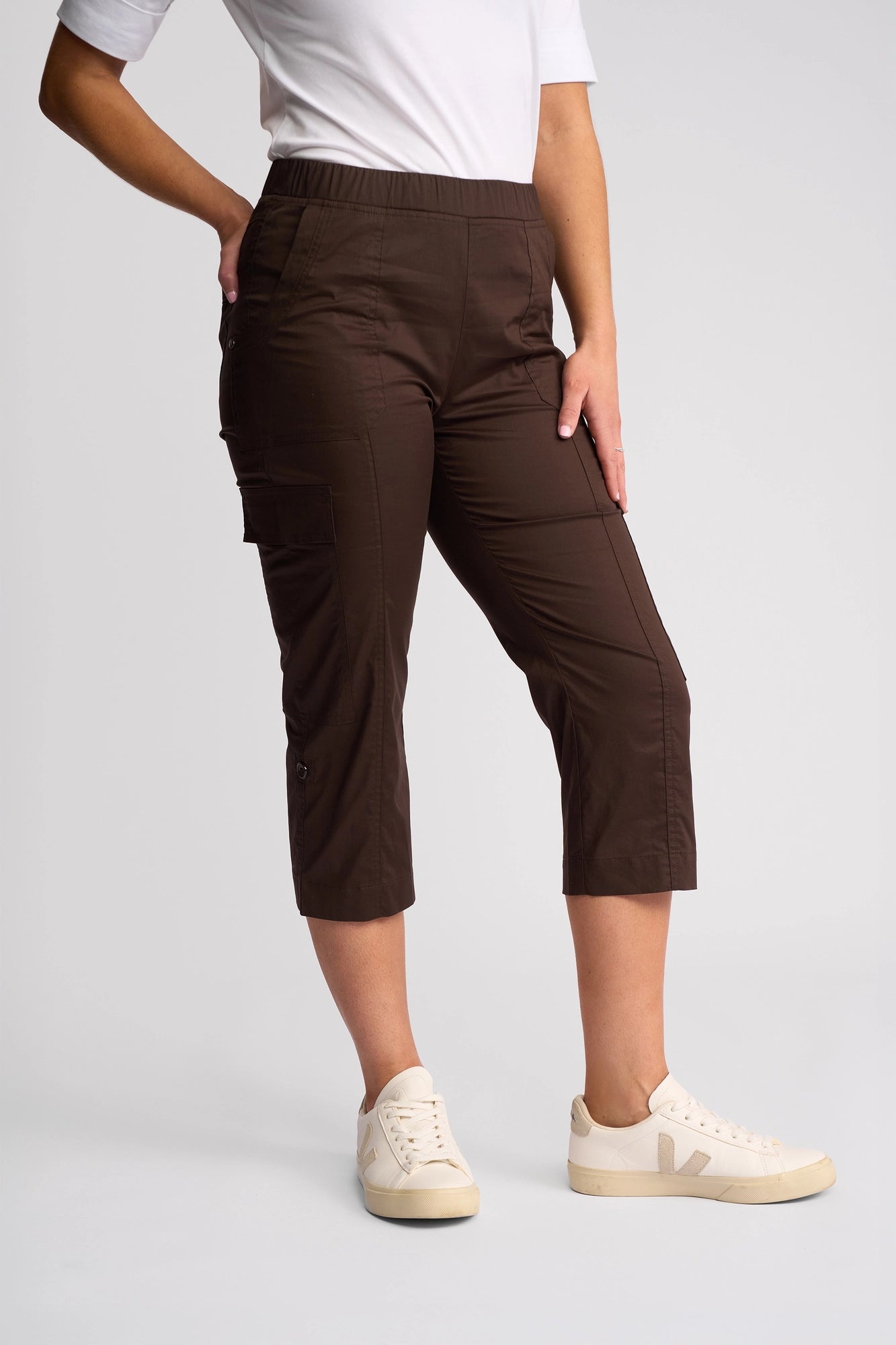 A person wearing a white short-sleeve shirt, Vassalli Wave Pant in Chocolate with side pockets and adjustable hems, and white sneakers, stands against a plain light-gray background.
