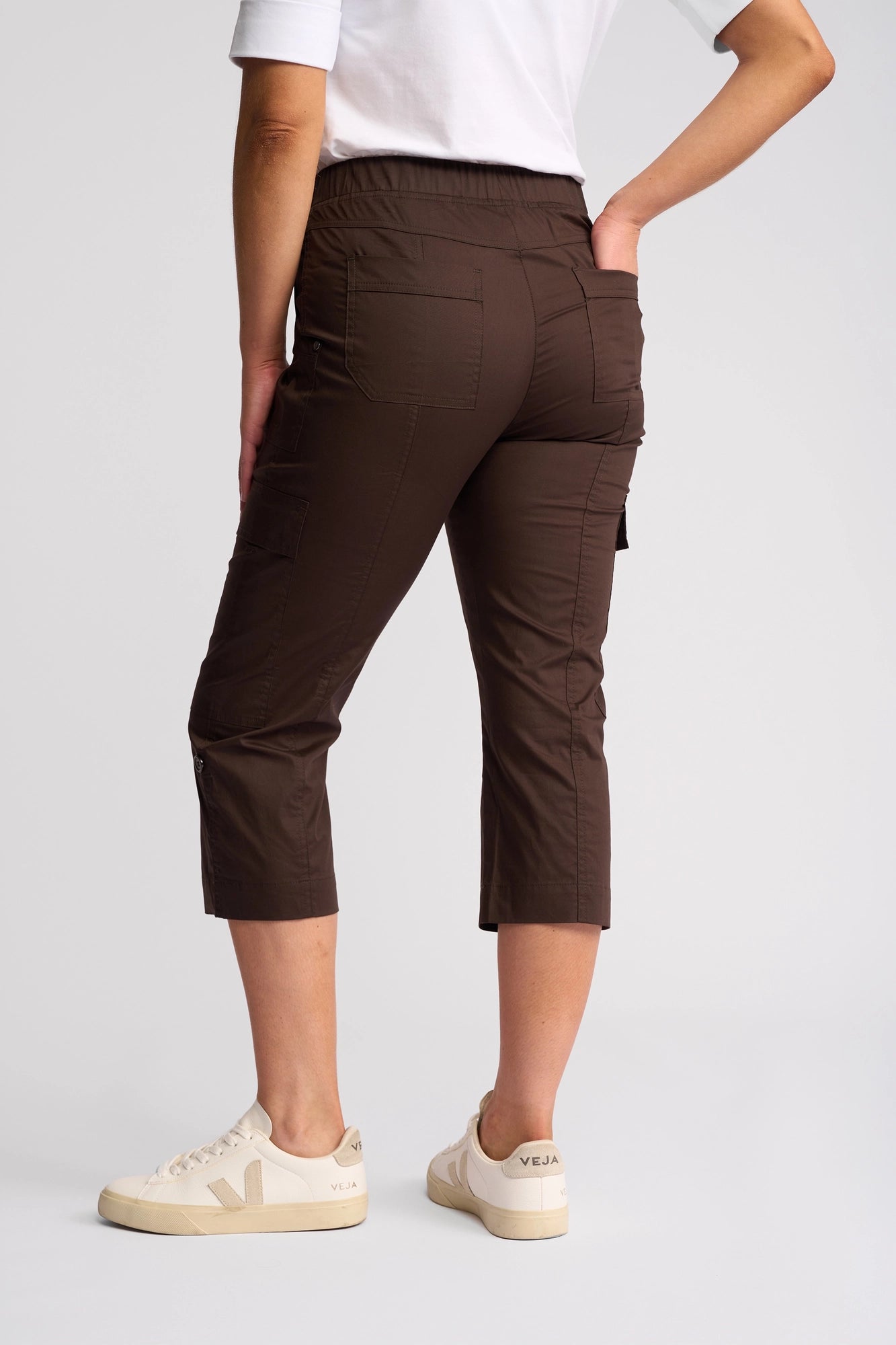 A person wearing a white t-shirt, Vassalli Wave Pant in Chocolate with an adjustable hem, and beige Veja sneakers stands facing away with one hand on their hip against a plain background.
