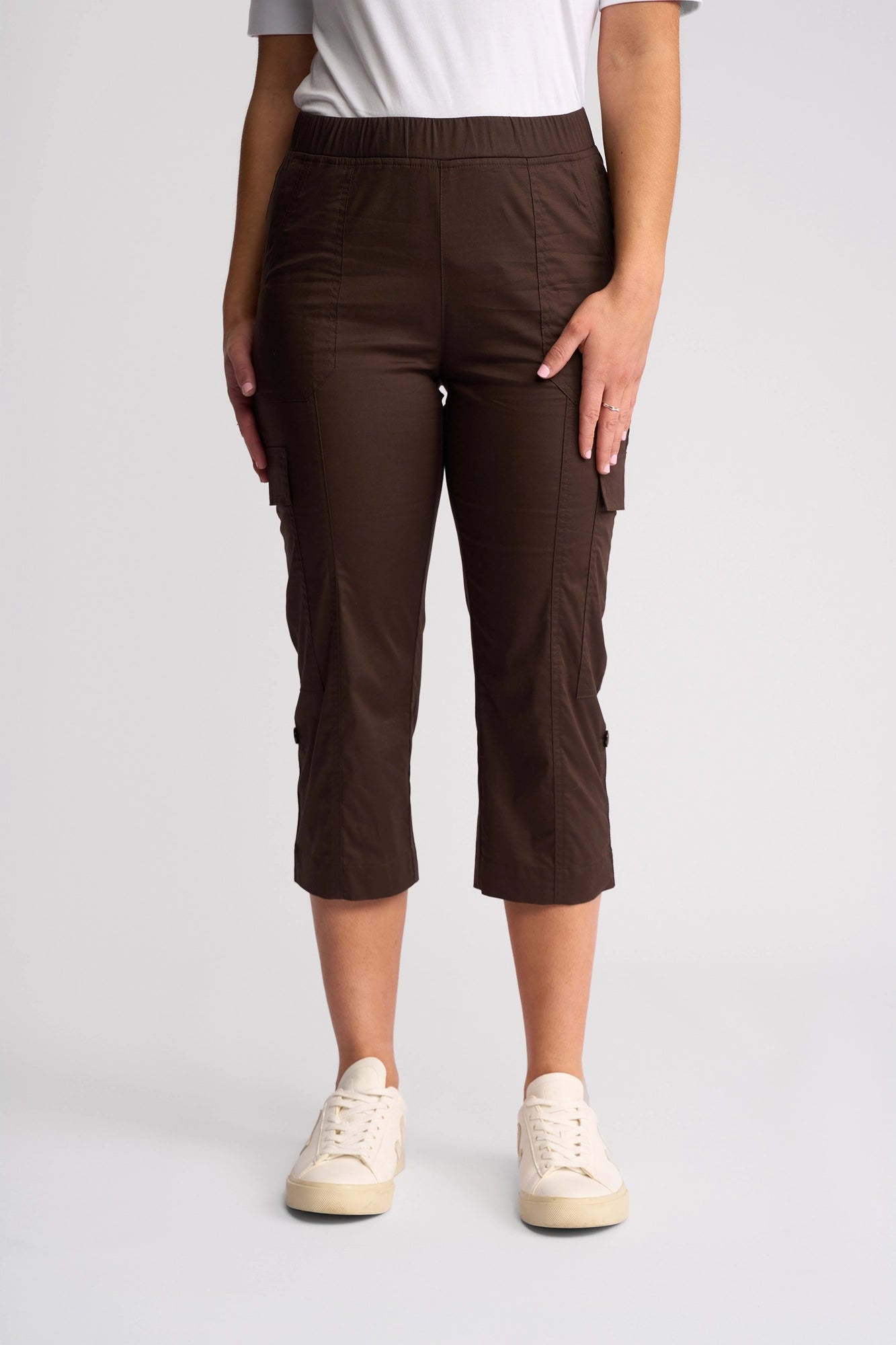 A person wearing Vassalli’s Wave Pant in Chocolate—a pair of brown stretch cotton cargo pants with an adjustable hem—paired with white sneakers, stands against a plain light background. The upper body is partially visible, featuring a white or light grey shirt.