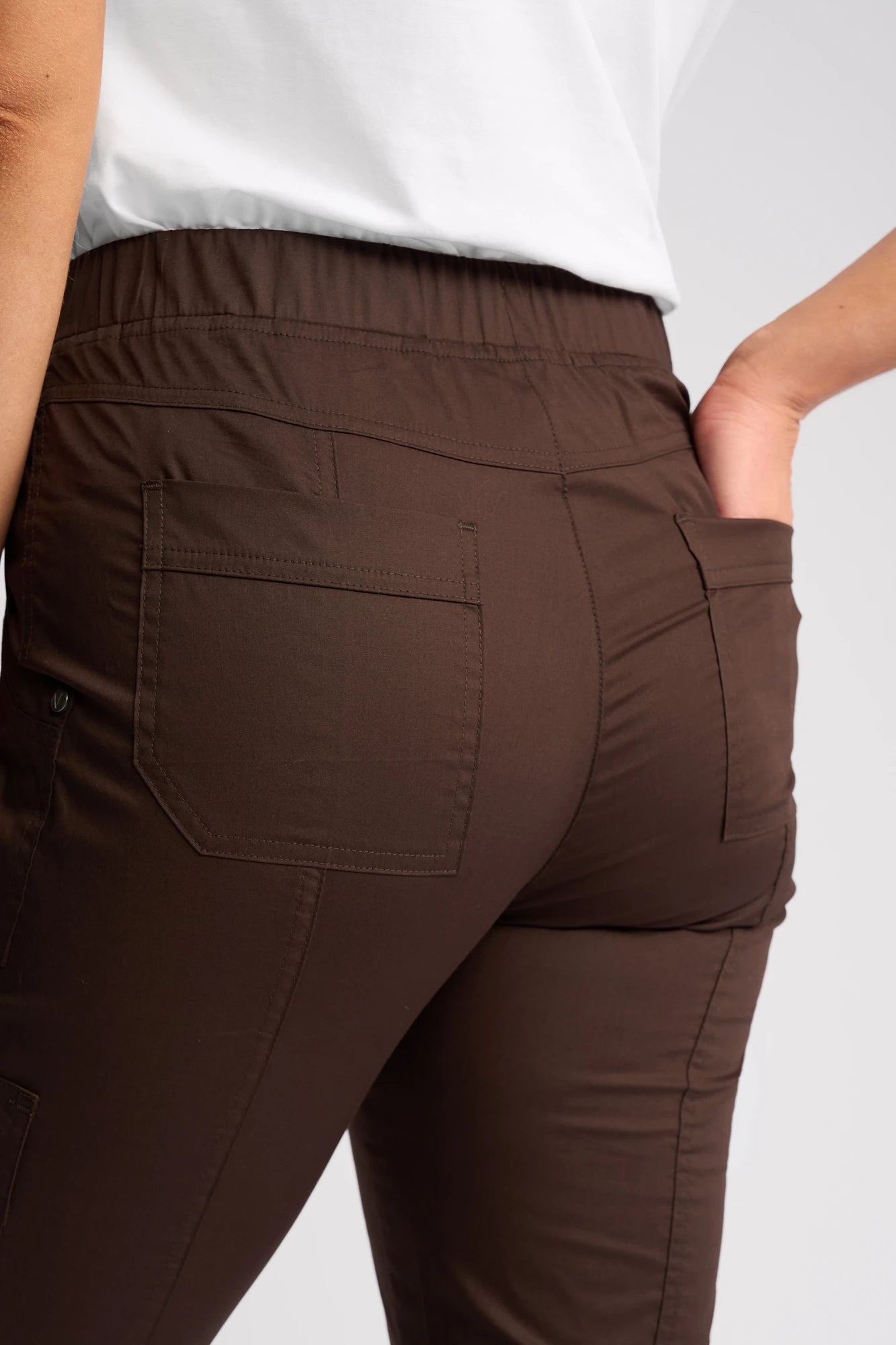 A person is shown from the waist down wearing Vassalli’s Wave Pant in Chocolate with a white shirt. The pants feature back pockets, and the person’s right hand is placed in one of the pockets.