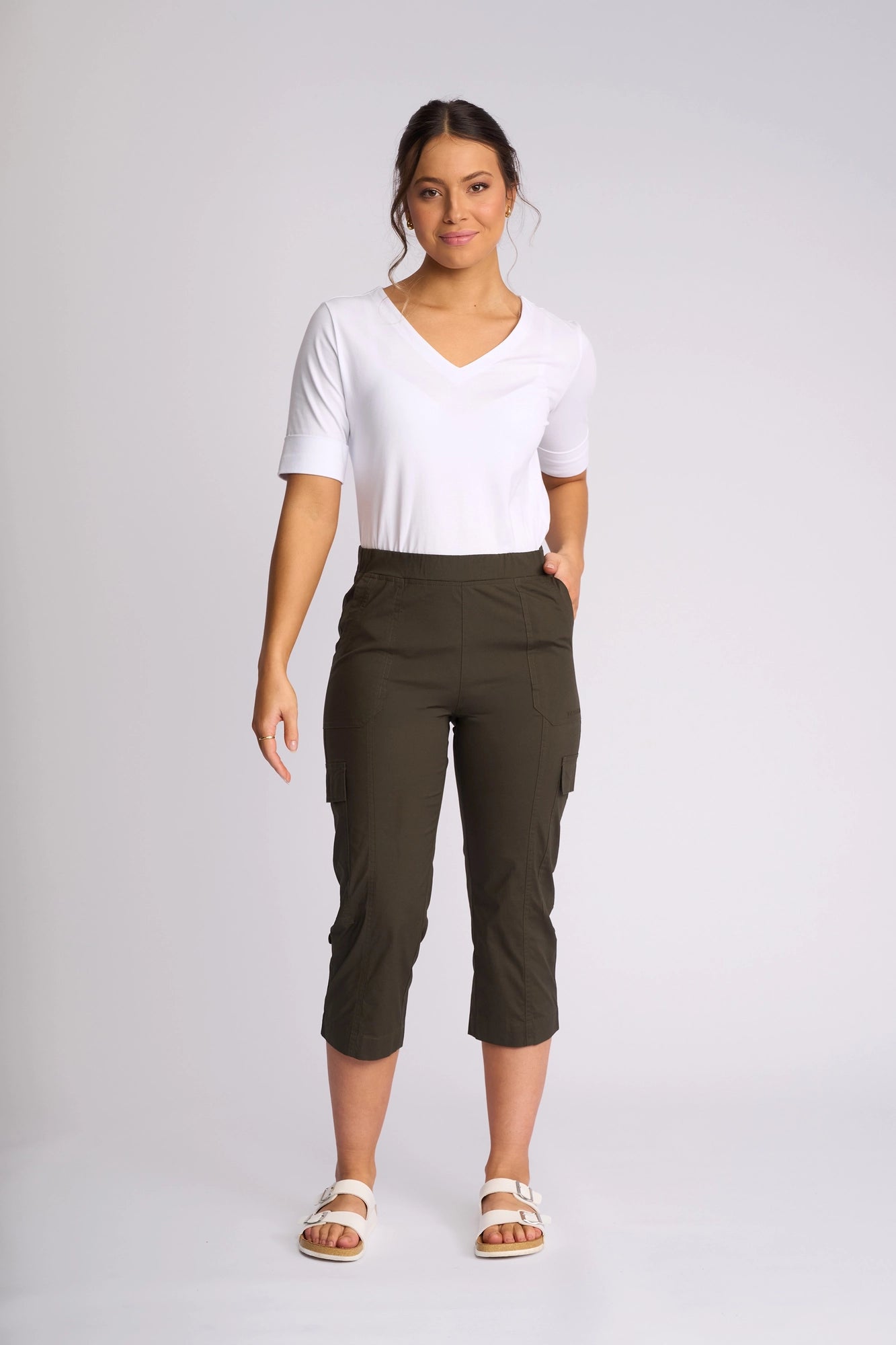 A woman stands facing forward, wearing a white V-neck top, the Vassalli Wave Pant in khaki, and white sandals, against a plain light background.