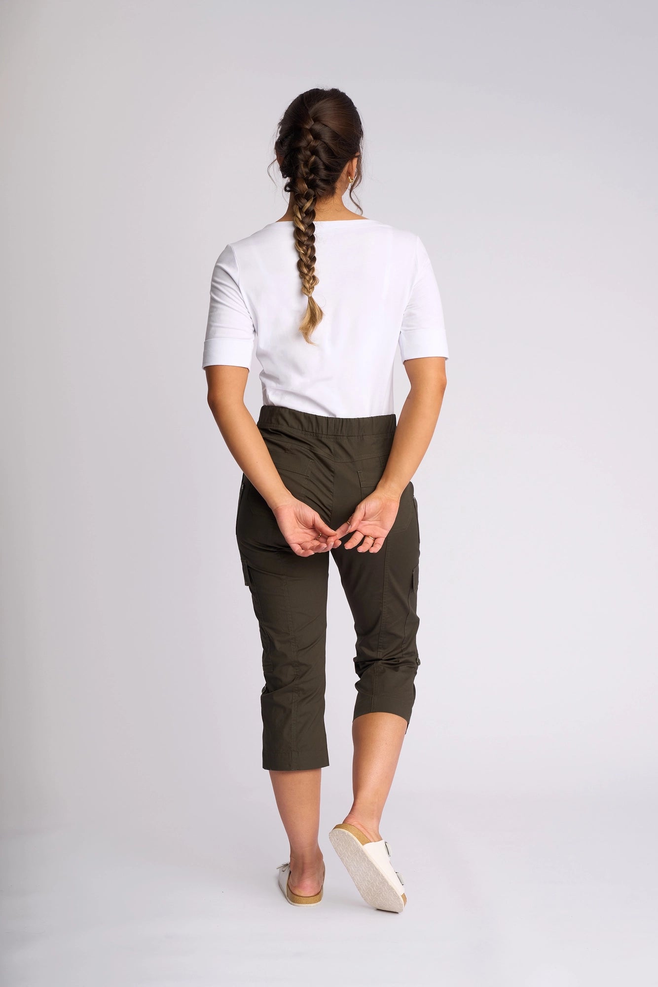 A woman with a long braid stands facing away, wearing a white shirt and the Vassalli Wave Pant in Khaki, featuring adjustable hems, paired with white slip-on shoes. Her hands are clasped behind her back against a plain light background.
