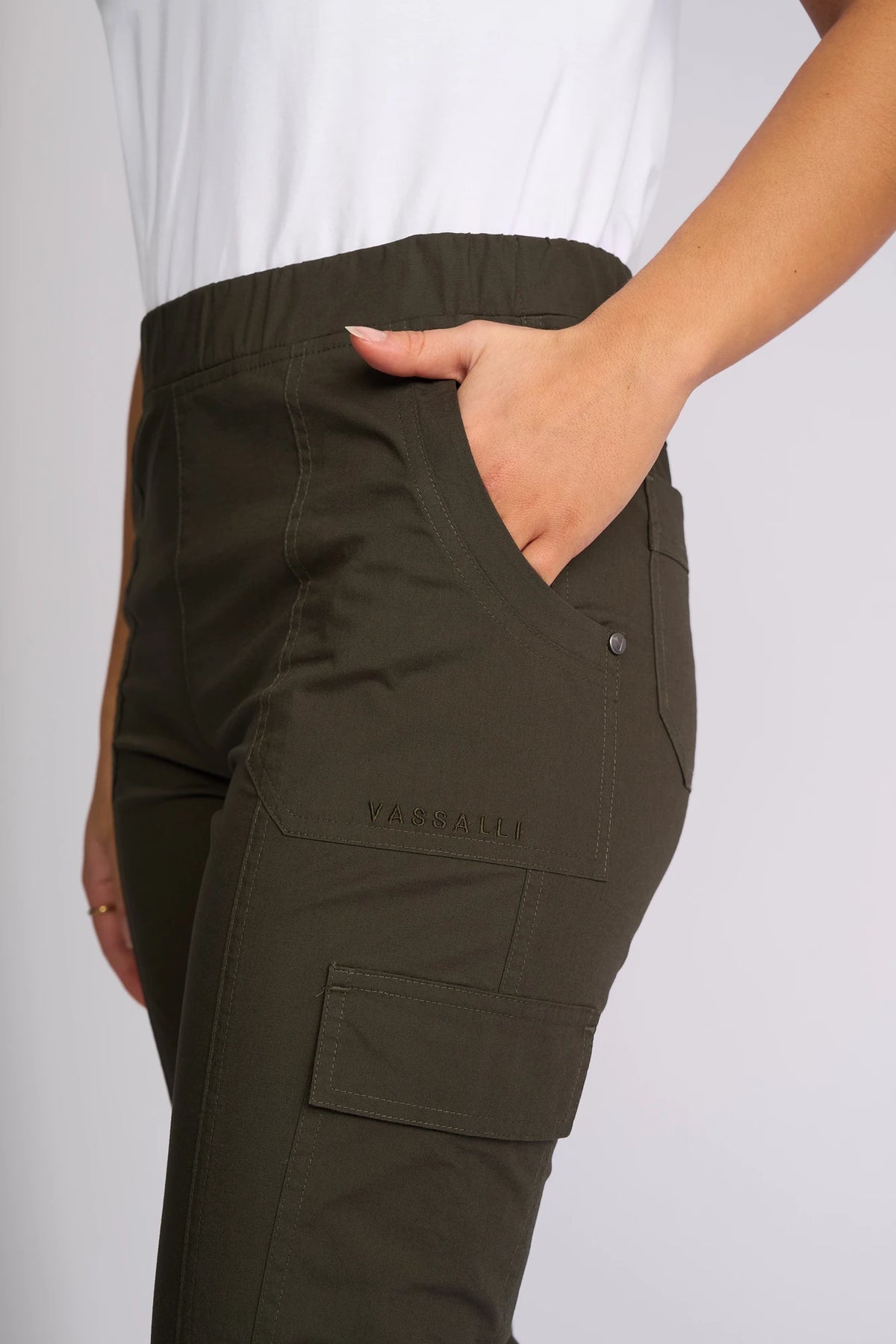 A person wearing the Vassalli Wave Pant in khaki—a stretch cotton cargo pant with stitching details, a flap pocket, adjustable hems, and “VASSALLI” text embroidered near the pocket—stands with one hand in their pocket and pairs it with a white top.