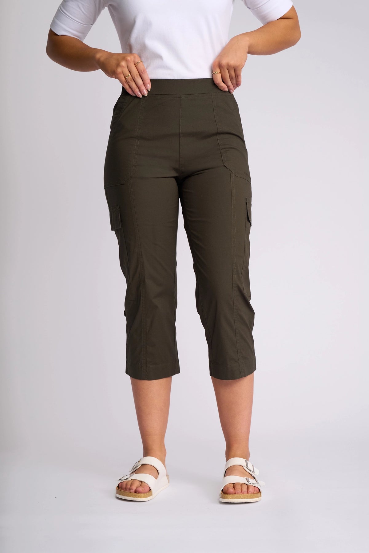 A person wearing the Vassalli Wave Pant in Khaki—crafted from stretch cotton with adjustable hems—paired with a white short-sleeve top and white sandals stands against a plain light background.