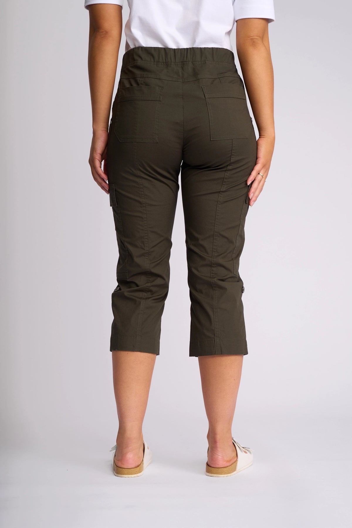 A person stands facing away, wearing the Vassalli Wave Pant in Khaki, featuring adjustable hems, paired with a white shirt and white slip-on shoes against a plain light background.
