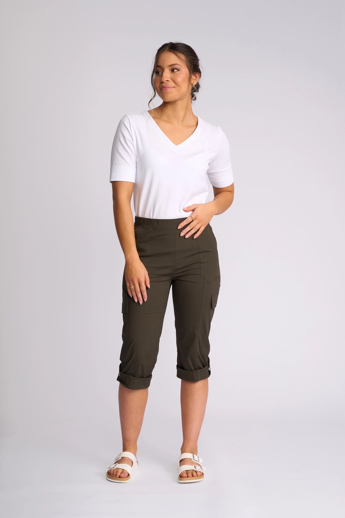 A woman stands in a studio wearing the Vassalli Wave Pant in Khaki, paired with a white V-neck shirt and white sandals. Her hair is tied back as she looks to the side with one hand on her waist.
