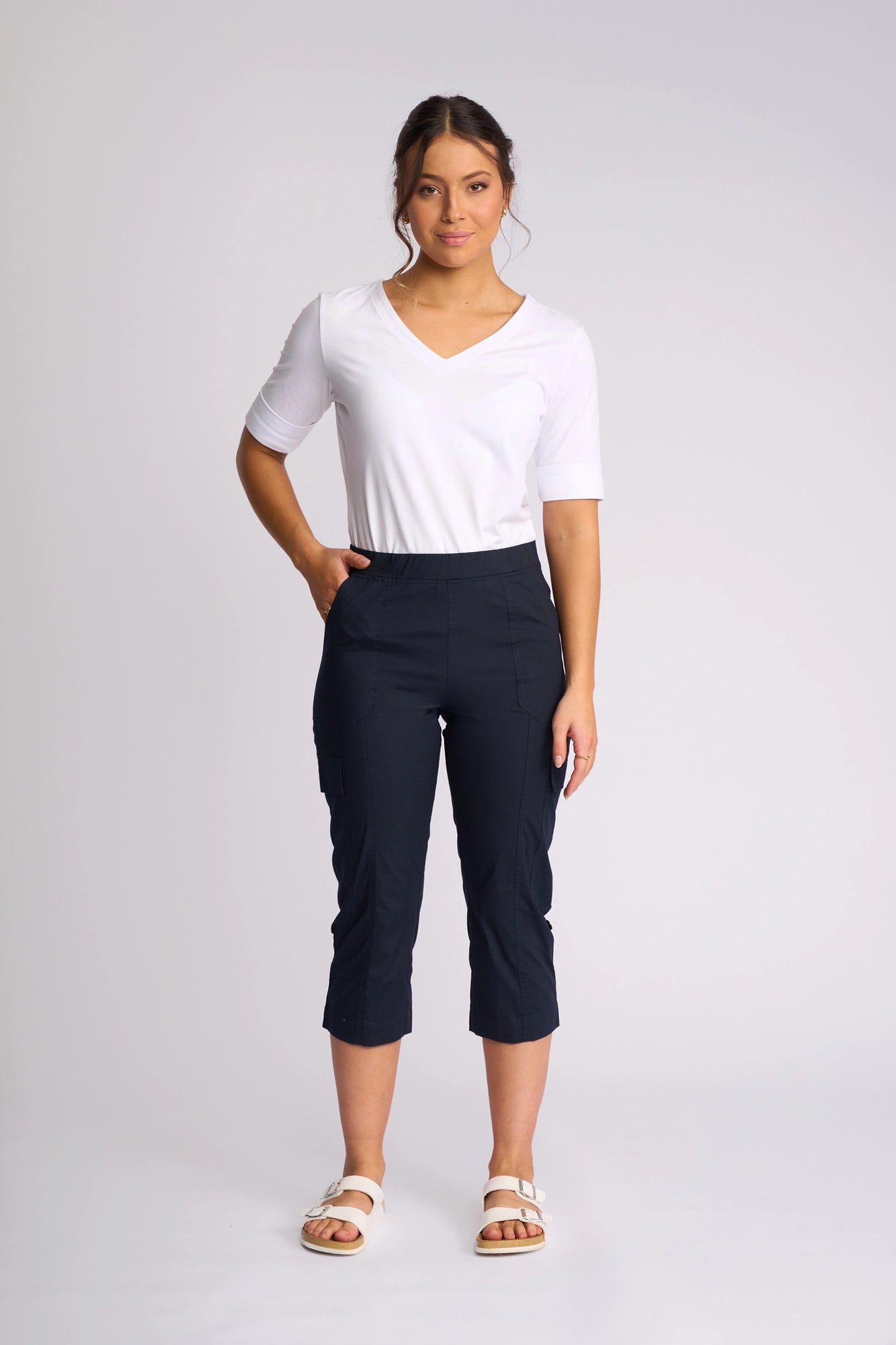 A woman stands against a plain background, facing forward with one hand in her pocket. She is wearing the Vassalli Wave Pant in navy—a stretch cotton capri style with adjustable hems—paired with a white short-sleeve top and white sandals.