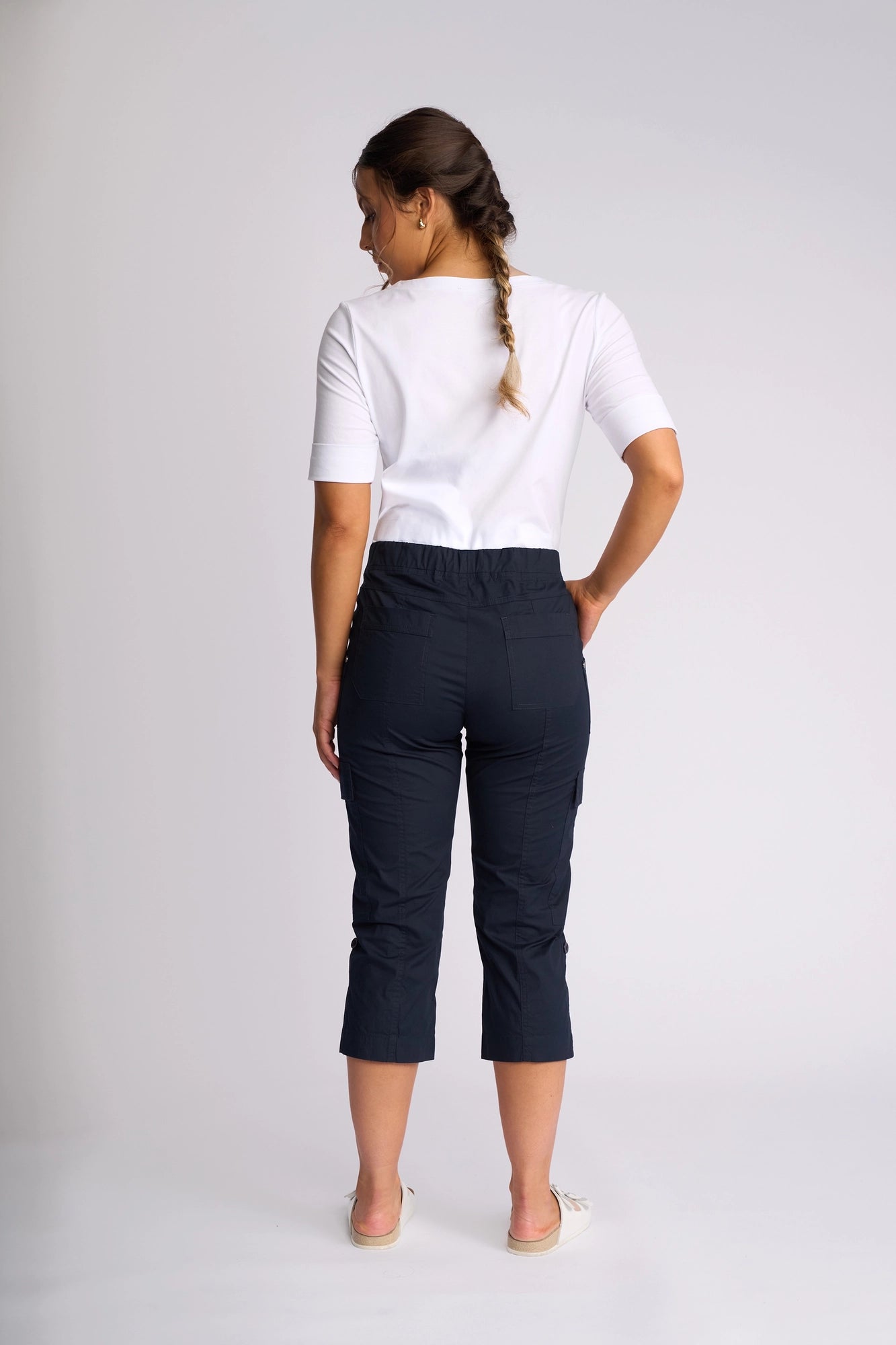 A woman with a braid stands facing away, wearing a white short-sleeve top and Vassalli’s Wave Pant in Navy, featuring stretch cotton fabric, pockets, and adjustable hems. She is barefoot on a plain light background.