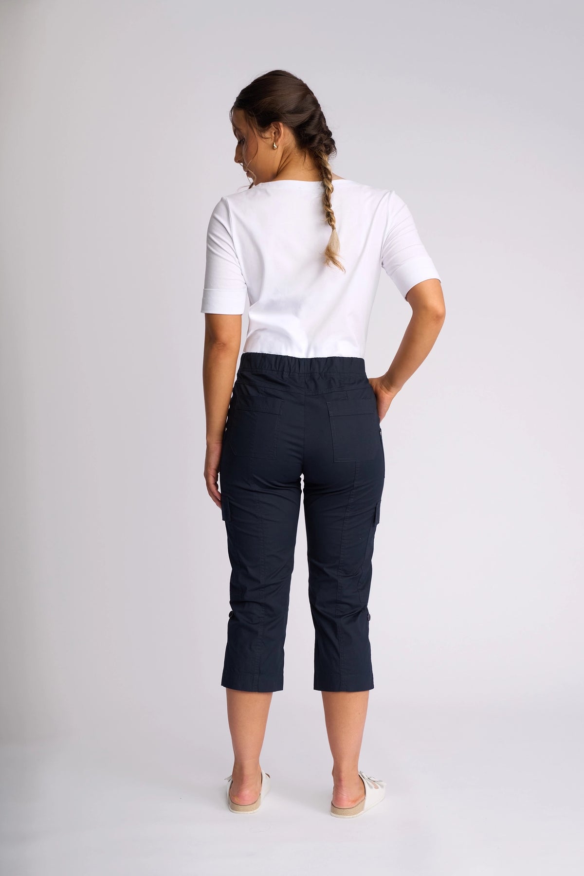 A woman with a braid stands facing away, wearing a white short-sleeve top and Vassalli’s Wave Pant in Navy, featuring stretch cotton fabric, pockets, and adjustable hems. She is barefoot on a plain light background.