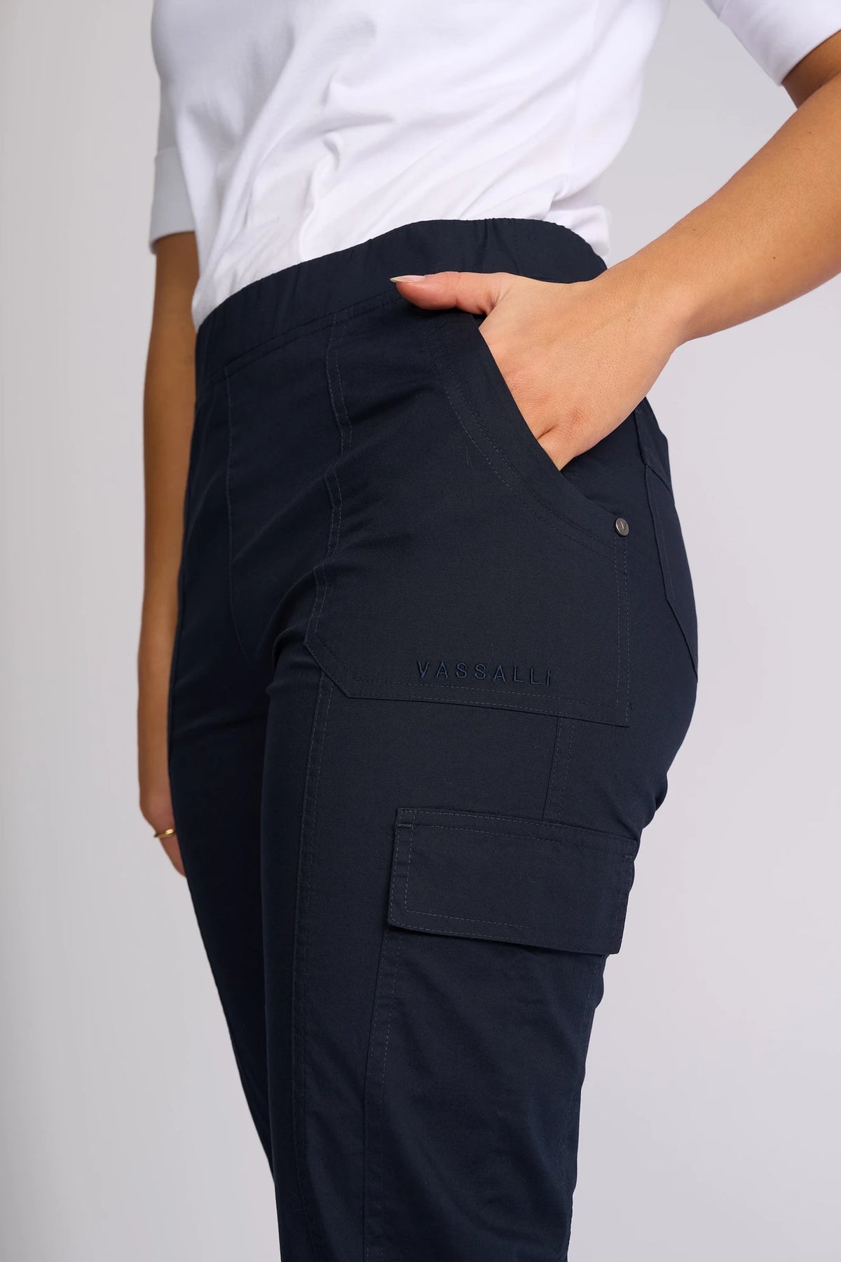 A person wearing the Vassalli Wave Pant in navy, made from stretch cotton and featuring VASSALLI embroidery on the pocket, stands with one hand in the pocket and pairs it with a white top.