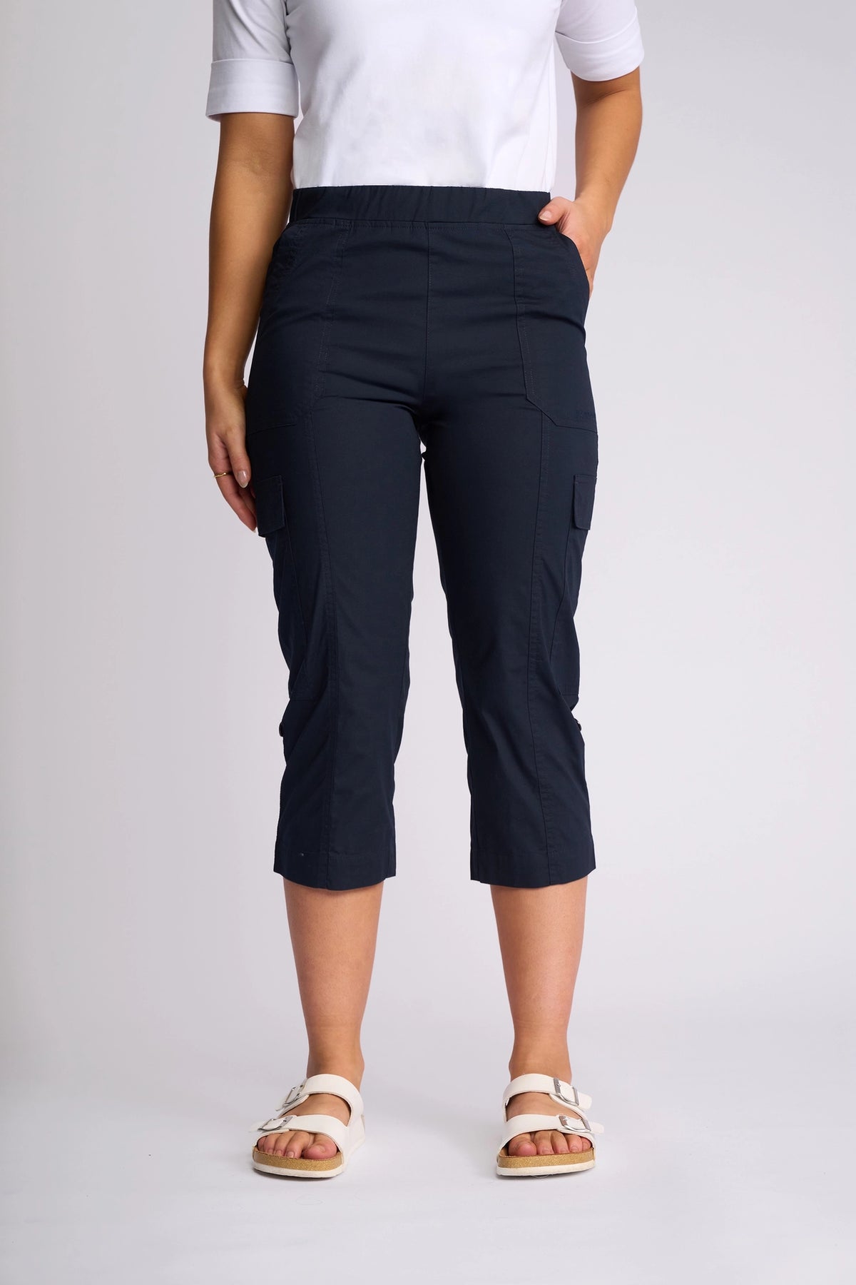 A person wearing a white top and the Vassalli Wave Pant in navy—a cropped cargo pant with adjustable hems made from stretch cotton—stands with one hand in a pocket against a plain light background.