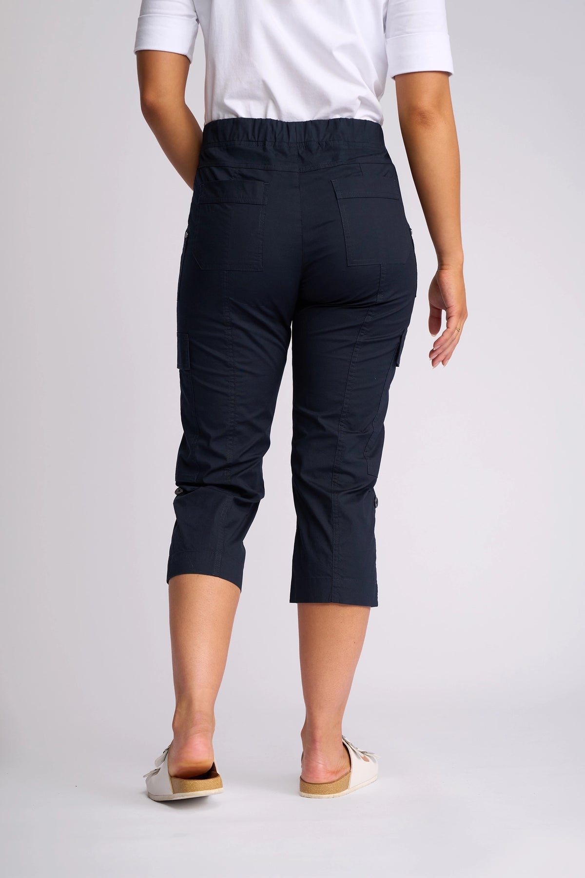 A person wearing a white shirt and the Vassalli Wave Pant in navy stands facing away, with adjustable hems on the cropped pants ending below the knee and light slip-on shoes.