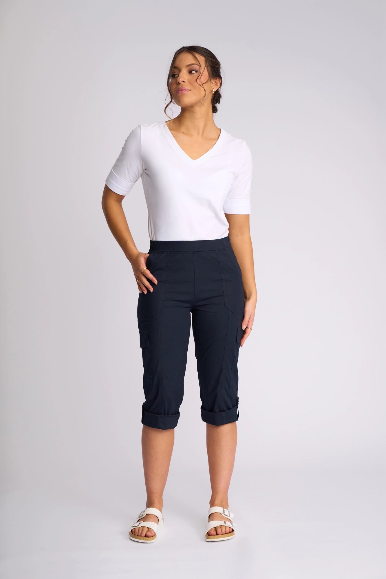 A woman stands against a plain background wearing the Vassalli Wave Pant in Navy, featuring stretch cotton fabric, cargo pockets, and adjustable hems. She pairs it with a white V-neck T-shirt and white sandals, posing with one hand on her hip while looking off to the side.