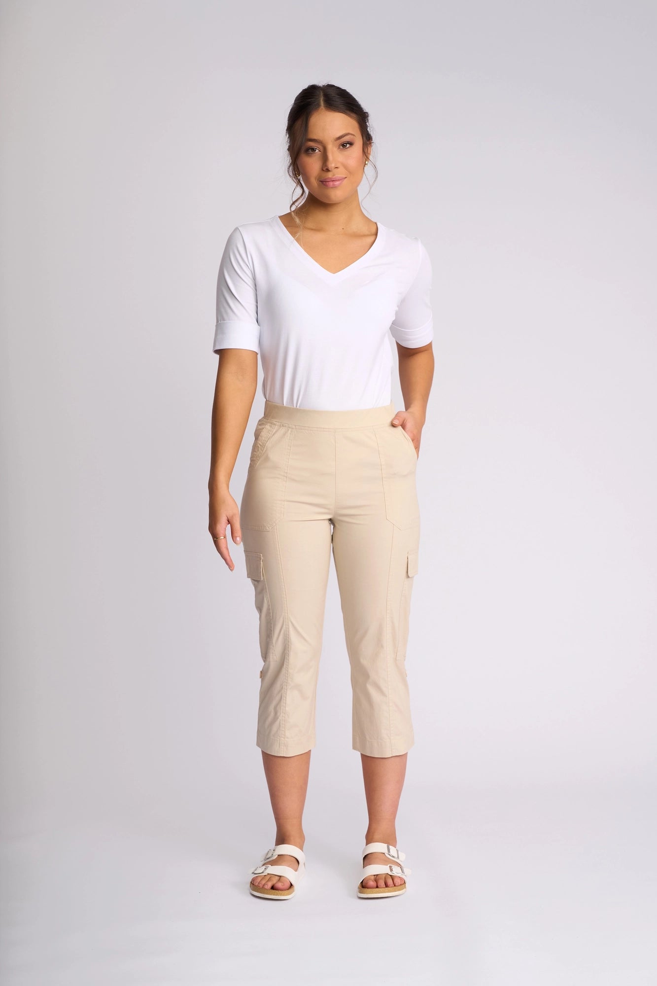 A woman stands against a plain background wearing a white short-sleeve V-neck top, the Vassalli Wave Pant in Sand—beige poplin cargo pants crafted from stretch cotton with adjustable hems—and white sandals, with one hand in her pocket.