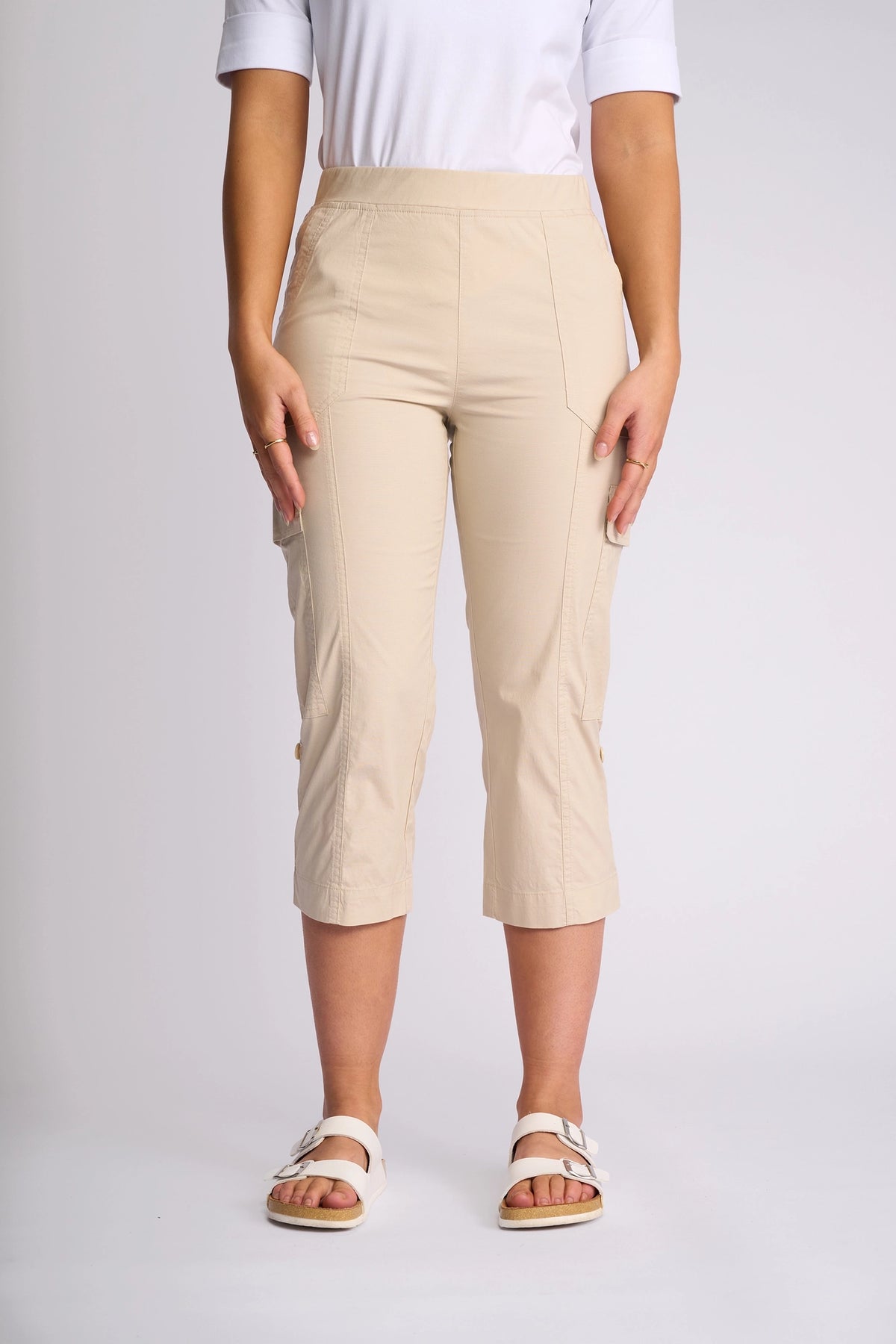 A person wearing a white shirt, Vassalli Wave Pant in Sand with adjustable hems, and white sandals stands against a plain, light background. The focus is on the clothing and lower body, with hands resting by the sides.