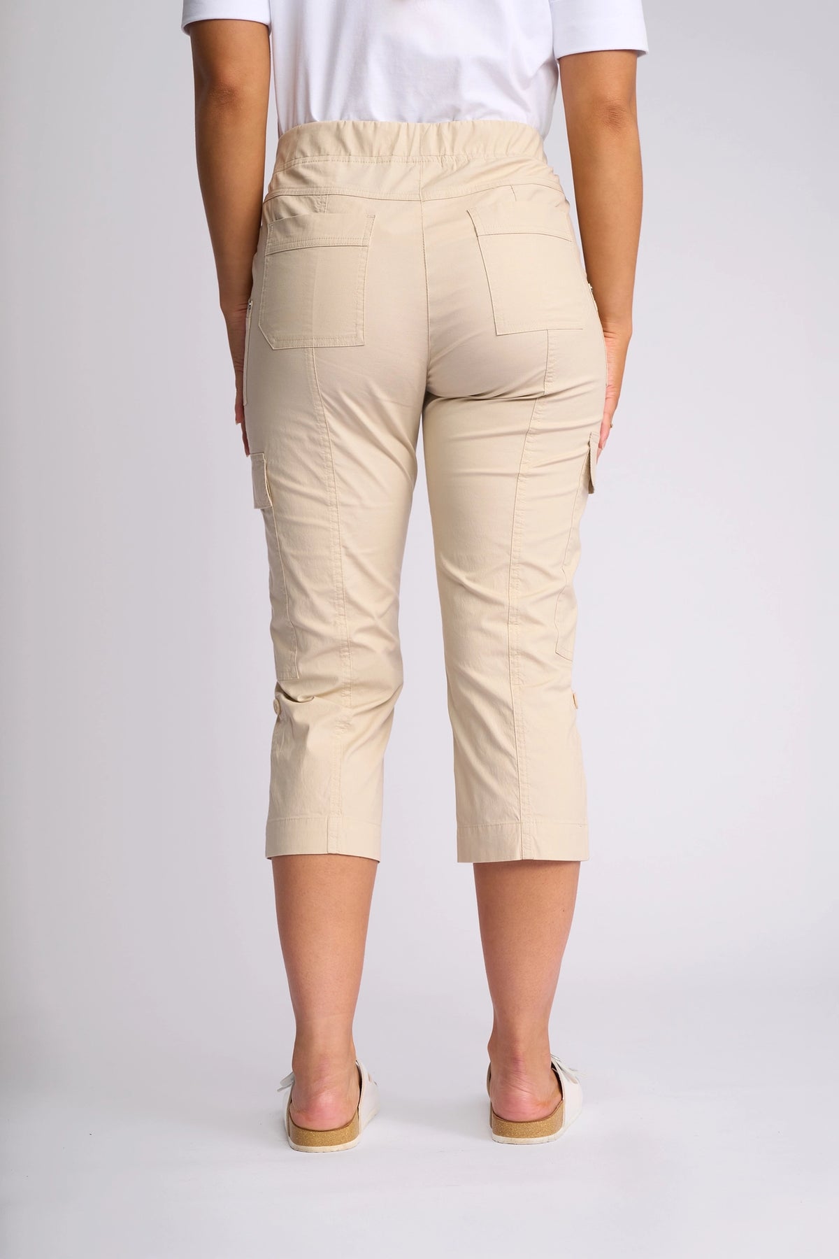 A person is seen from behind, standing against a plain background, wearing the Vassalli Wave Pant in Sand—a pair of beige poplin cargo pants with pockets and adjustable hems—paired with a white shirt and light-colored slip-on shoes.