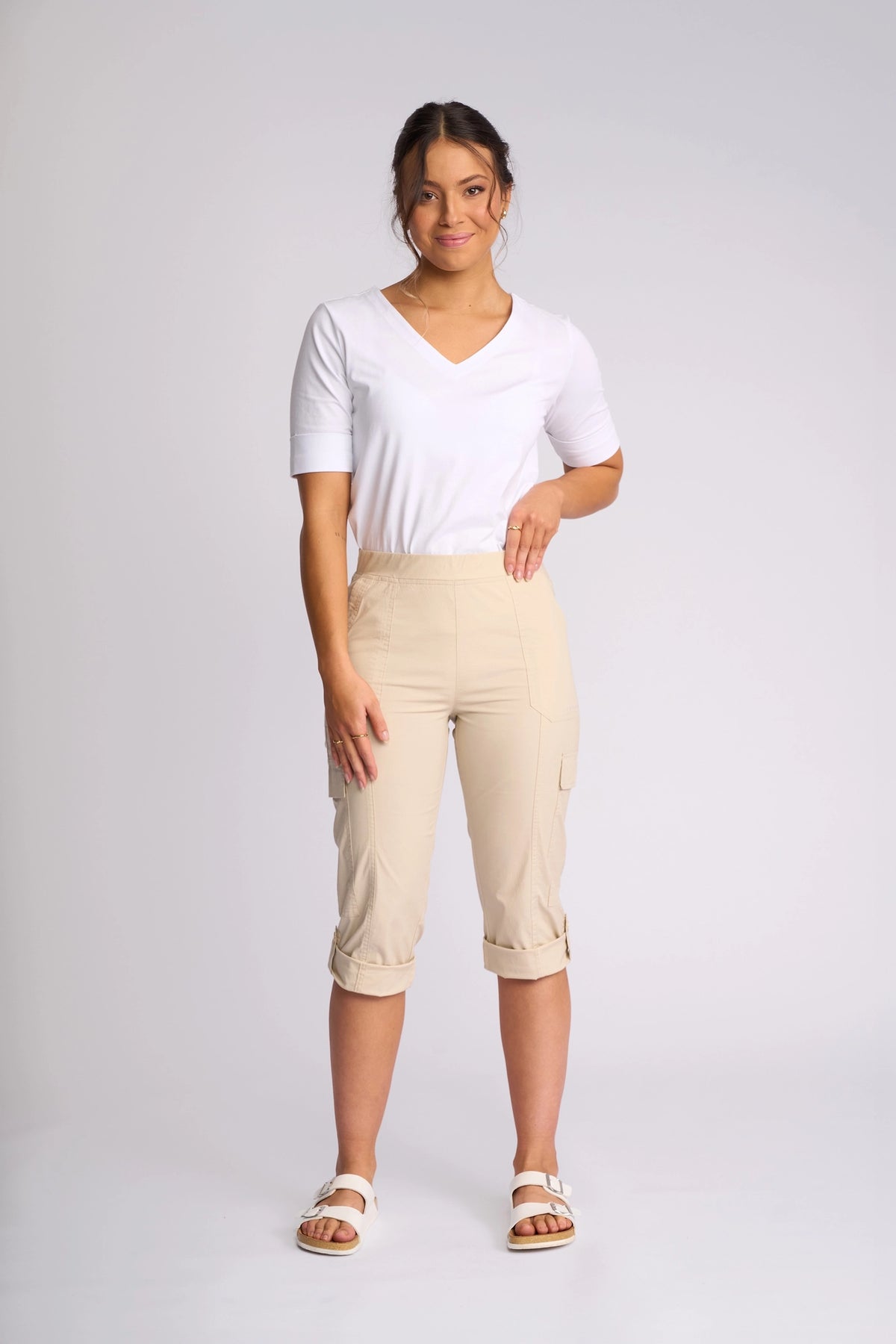 Smiling with one hand on her hip against a plain light background, the woman stands in a studio wearing a white short-sleeve top, the Vassalli Wave Pant in Sand—beige poplin cargo pants with adjustable hems crafted from stretch cotton—and white sandals.