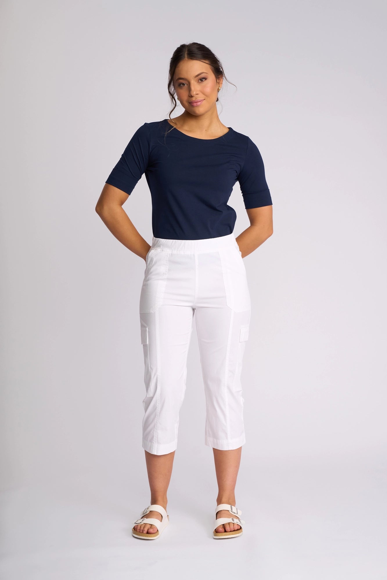 A woman stands against a light background wearing a navy blue short-sleeve top and the Vassalli Wave Pant in white with an adjustable hem, paired with white sandals, her hands behind her back.