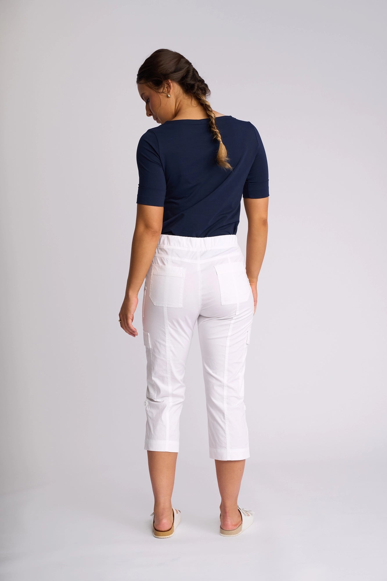 A woman with a braid, wearing a navy blue top and the Vassalli Wave Pant in white with an adjustable hem, stands barefoot facing away and looking down slightly. The background is plain and light-colored.