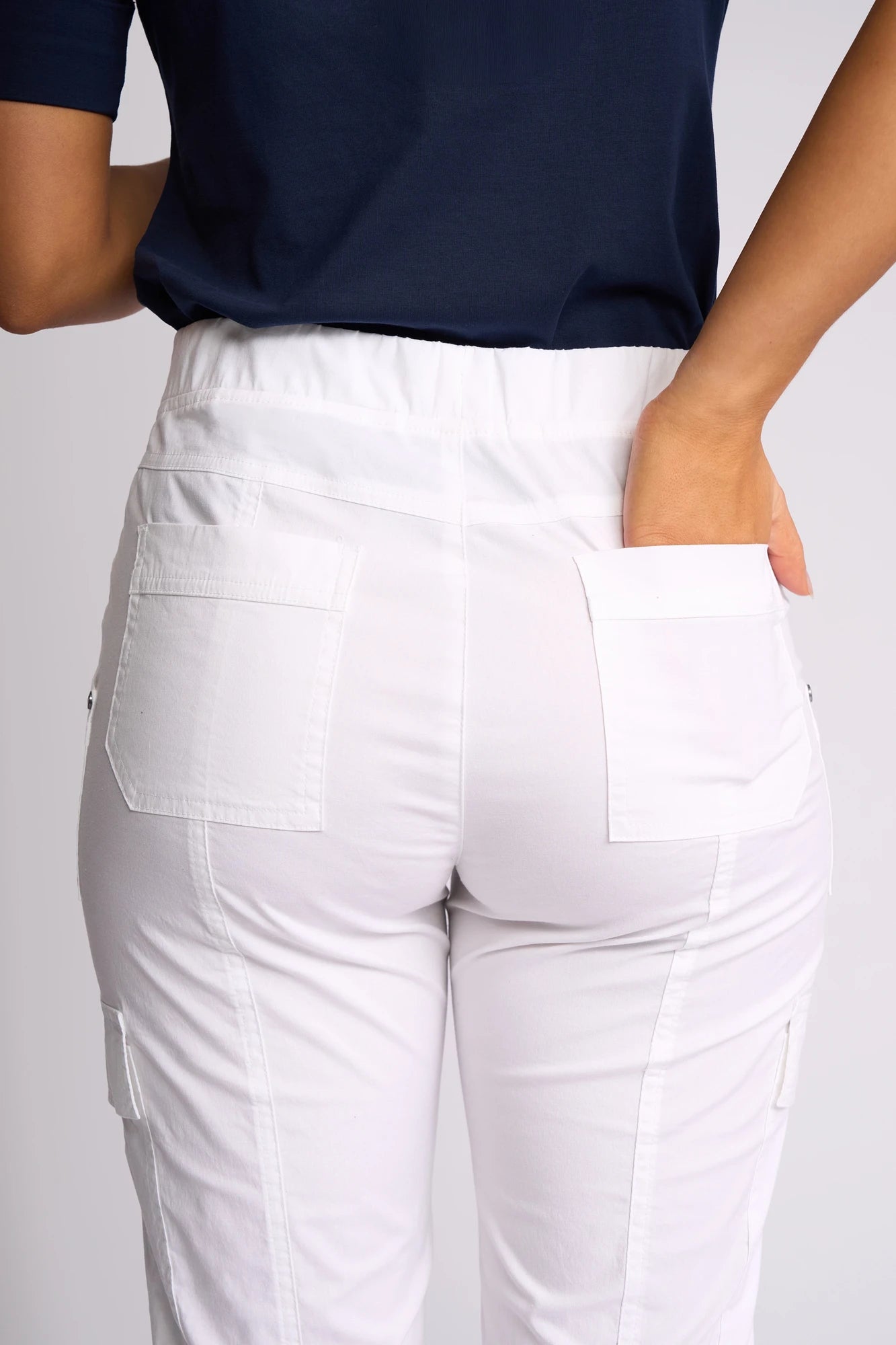A person is shown from the back wearing a navy top and Vassalli Wave Pant in White, featuring patch pockets and an adjustable hem for added versatility; one hand is placed in the back pocket.