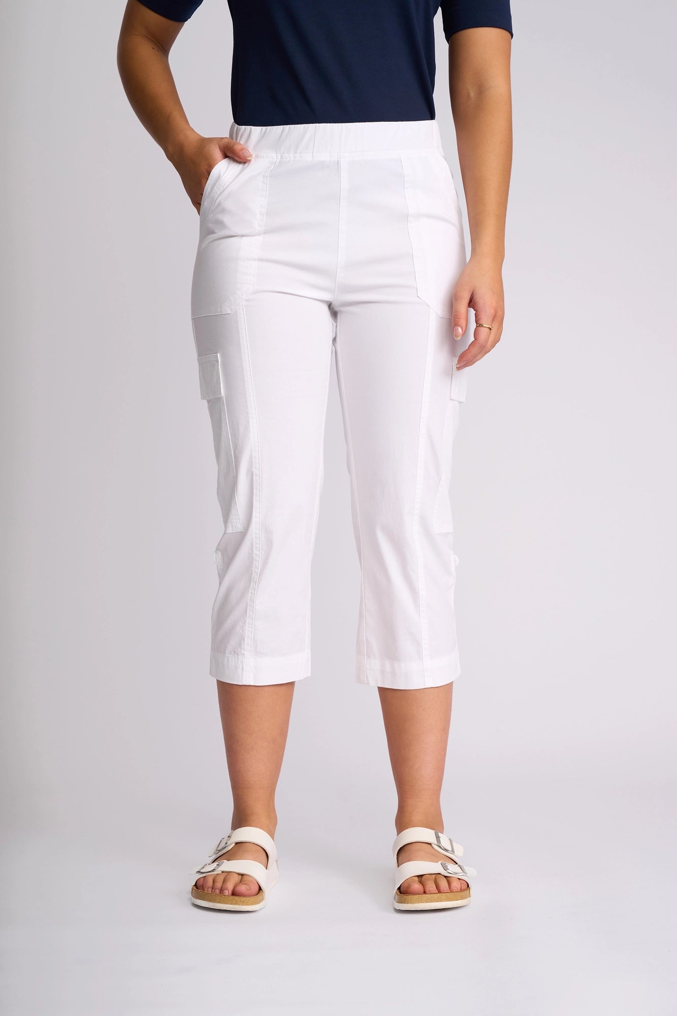 A person wearing a navy short-sleeve shirt, Vassalli Wave Pant in white with pockets and an adjustable hem, and white sandals stands against a plain background.