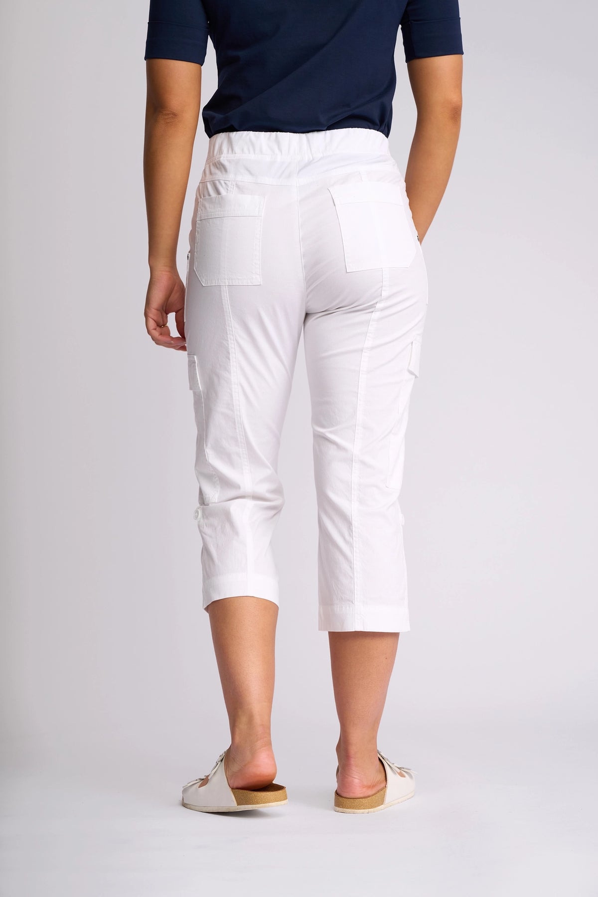A person is shown from the back, standing against a plain light background, wearing the Vassalli Wave Pant in white—a cropped poplin cargo style with an adjustable hem—paired with a navy shirt and white slip-on shoes.