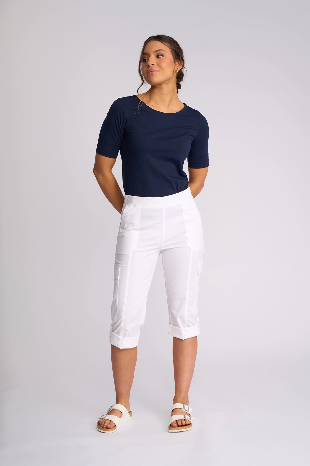 A woman stands against a plain background, wearing a navy blue short-sleeve top, the Vassalli Wave Pant in white with an adjustable hem, and white sandals. Her hands are behind her back as she looks off to the side.