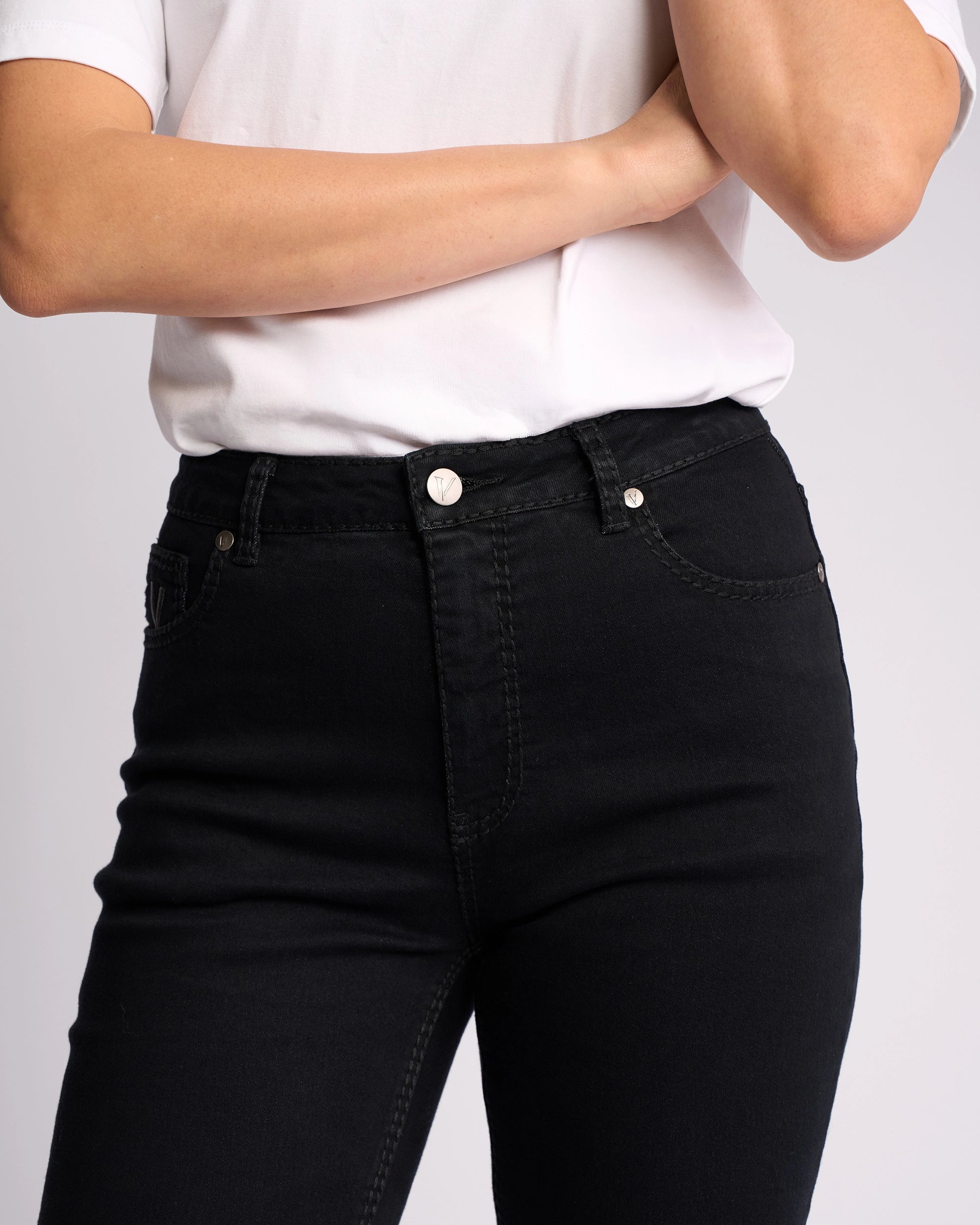A person models the Vassalli Lotus Jean in Black Denim, high-waisted and slim-leg style, paired with a white t-shirt, shown from shoulders to mid-thigh against a plain background with one arm crossed over their chest.