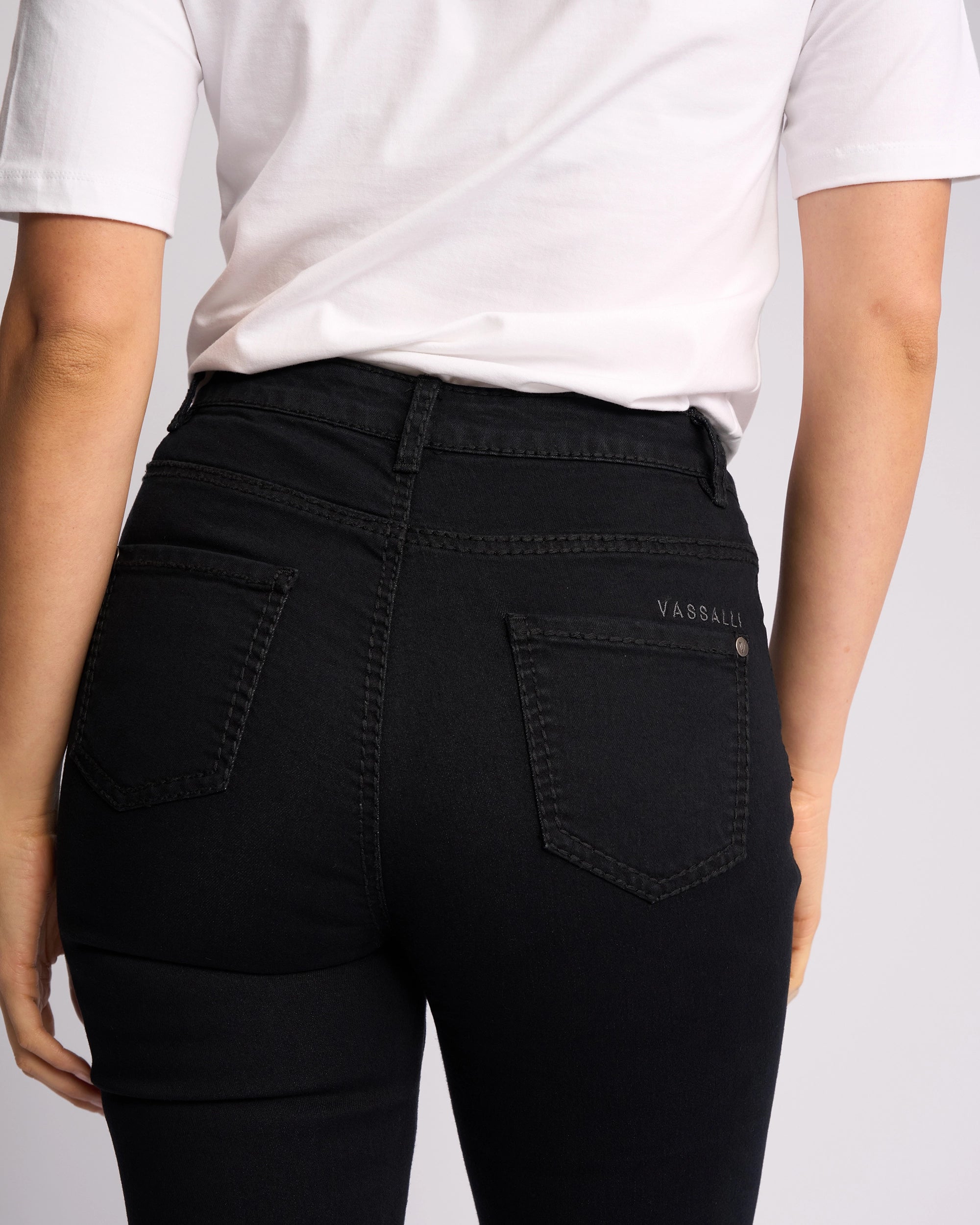 A person wearing a white t-shirt and Vassalli Lotus Jean - Black Denim, shown from the back. The women's jeans feature back pockets and a small "VASSALLI" label above the right pocket.
