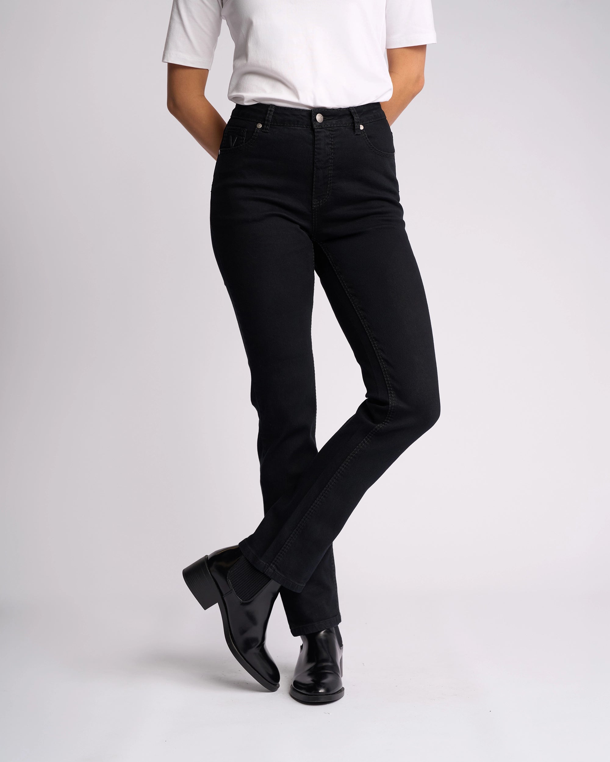 A person stands with arms behind their back and ankles crossed against a plain light background, wearing a white short-sleeve shirt, Vassalli Lotus Jean - Black Denim, and black ankle boots.