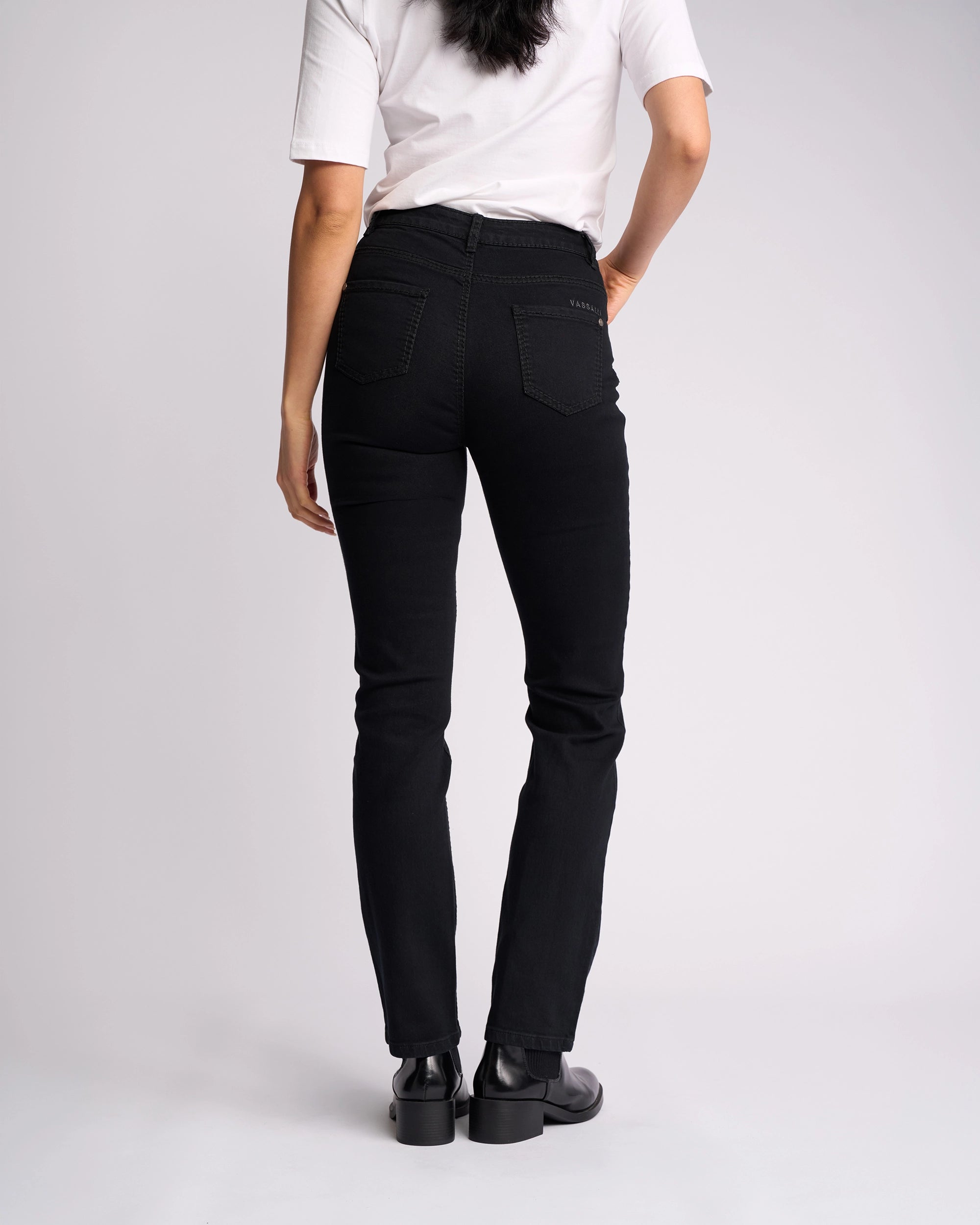 A person stands facing away from the camera, wearing a white short-sleeve shirt, Vassalli Lotus Jean - Black Denim, and black shoes against a plain light background.