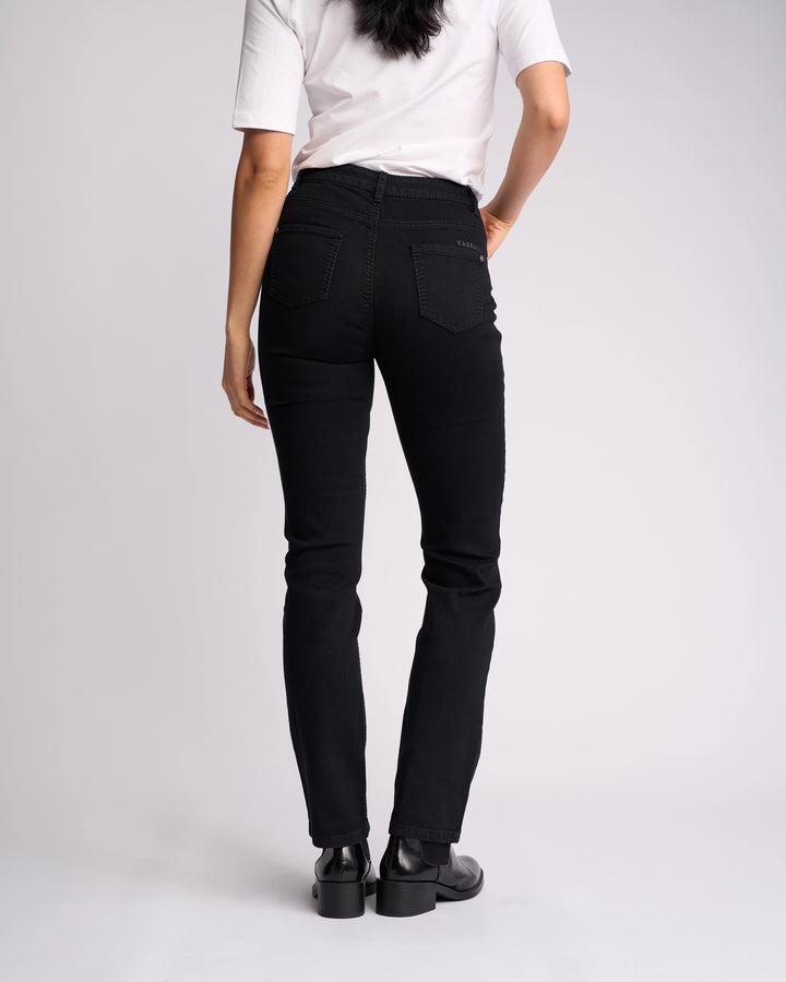 A person stands facing away from the camera, wearing a white short-sleeve shirt, Vassalli Lotus Jean - Black Denim, and black shoes against a plain light background.