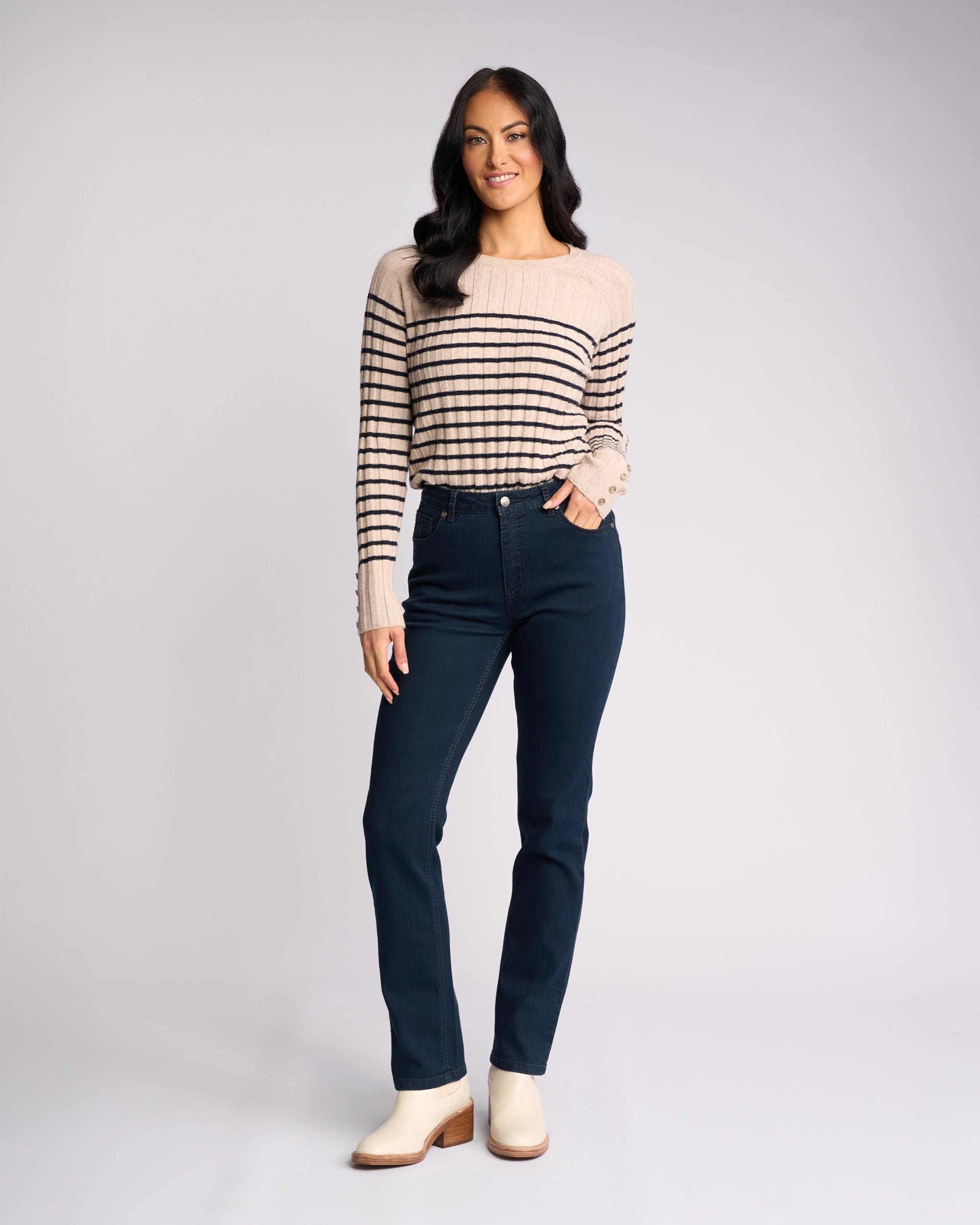 A woman with long dark hair smiles with one hand in her pocket, wearing the Vassalli Lotus Jean in Indigo Denim, a beige and black striped sweater, and light ankle boots against a plain light background.