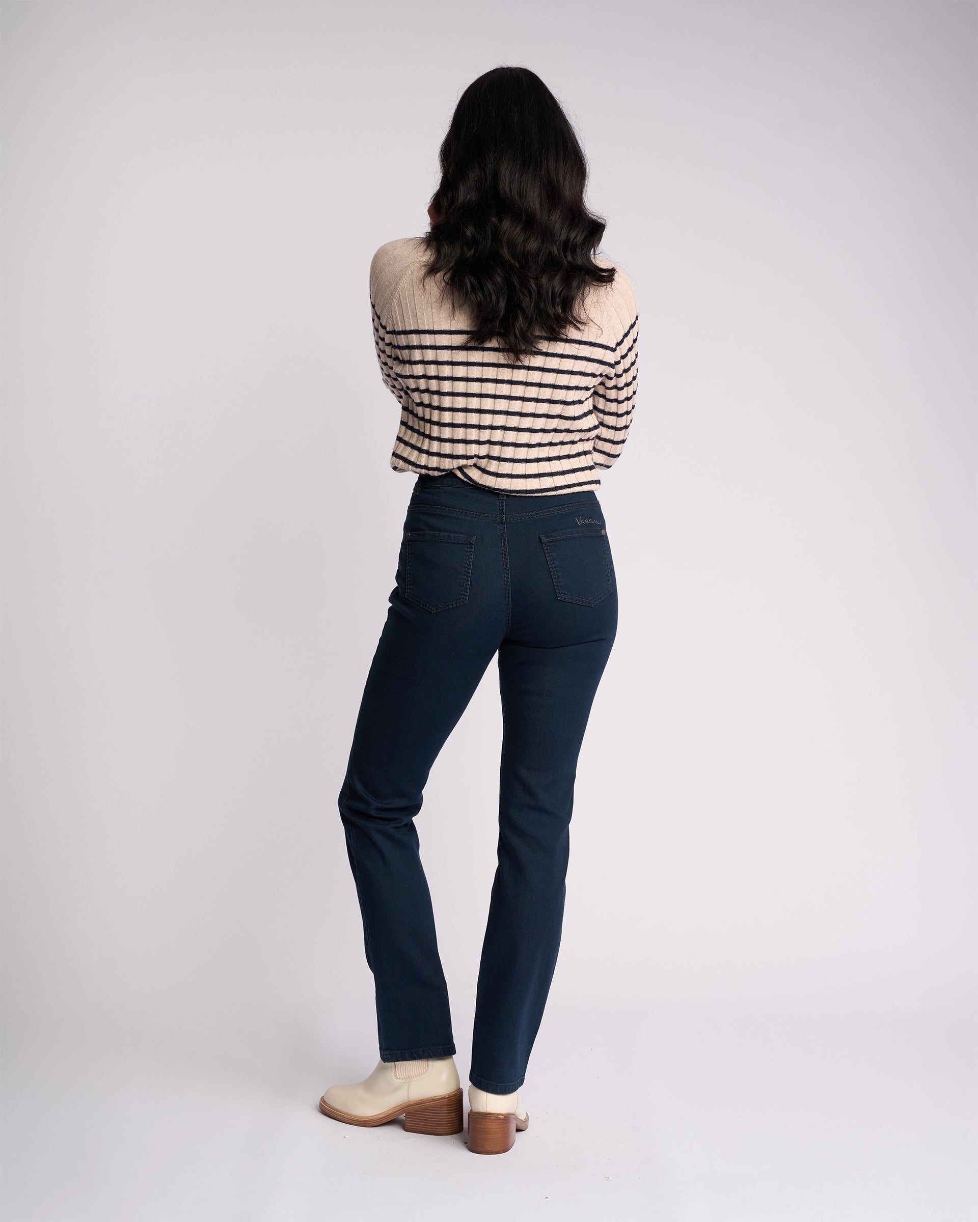 A woman with long dark hair, wearing a beige and black striped sweater, cream shoes, and Vassalli Lotus Jean in indigo denim, stands facing away from the camera against a plain white background.
