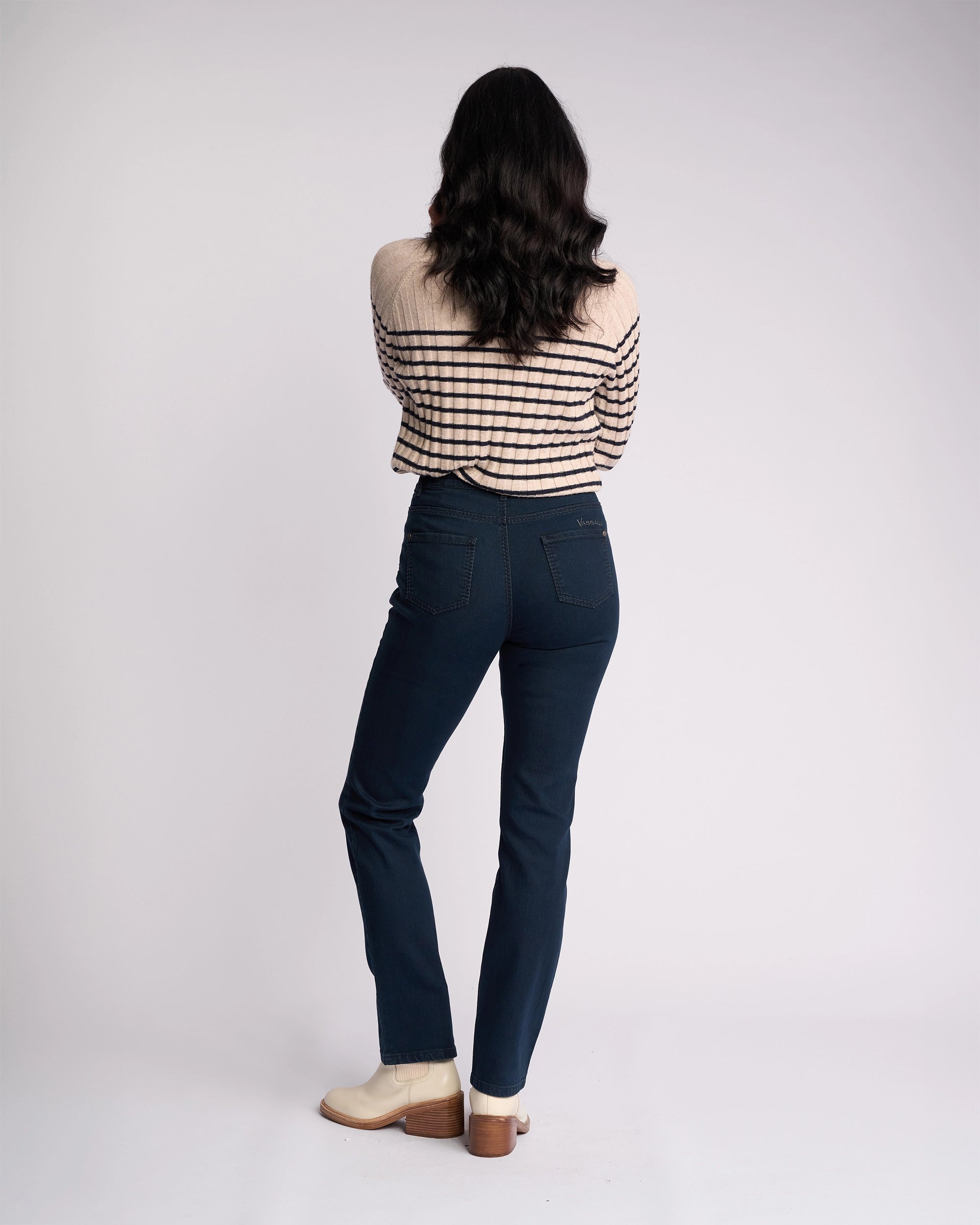 A woman with long dark hair, wearing a beige and black striped sweater, cream shoes, and Vassalli Lotus Jean in indigo denim, stands facing away from the camera against a plain white background.
