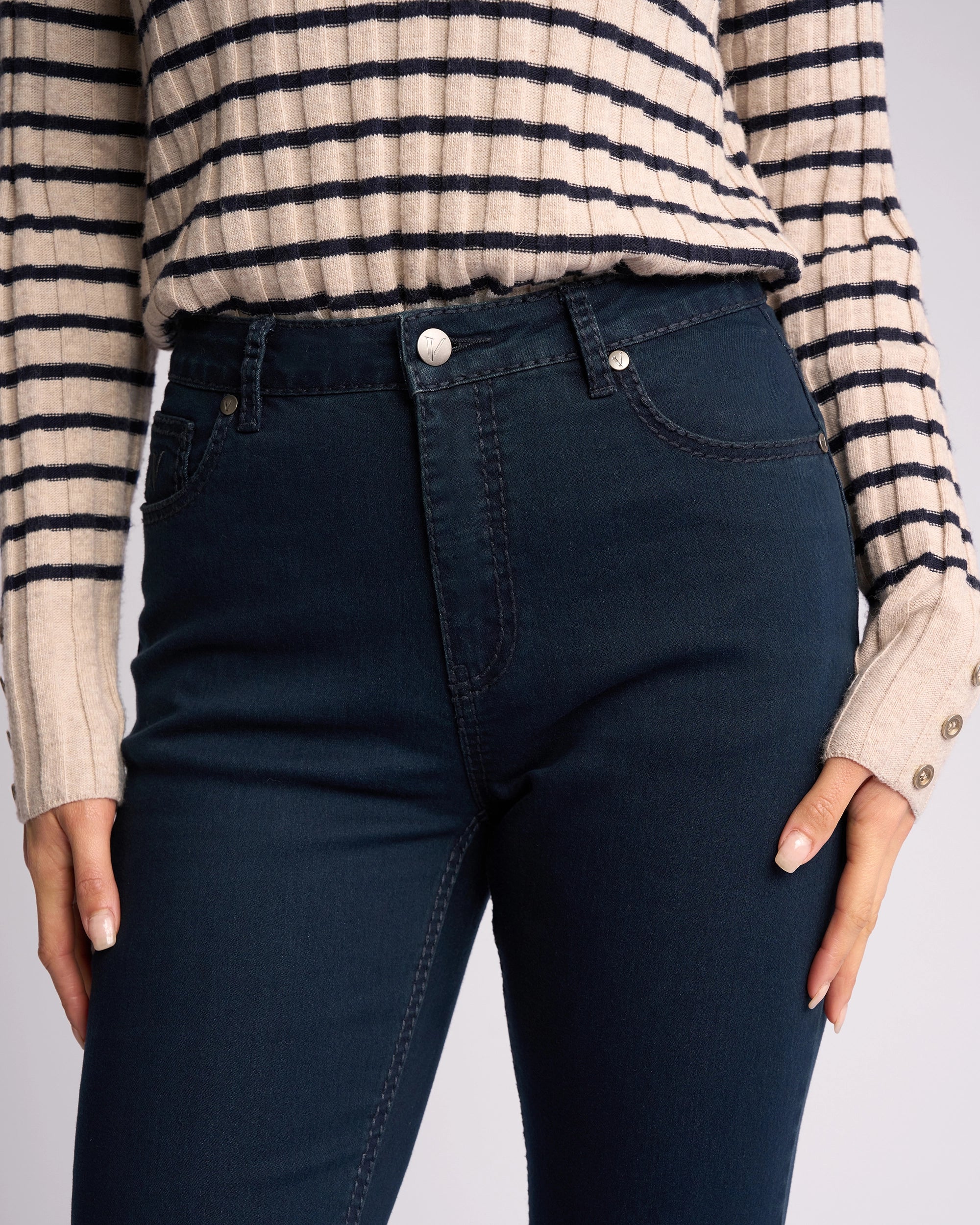 A person wearing a cream and navy striped long-sleeve sweater and Vassalli Lotus Jean - Indigo Denim, with their hands resting at their sides.